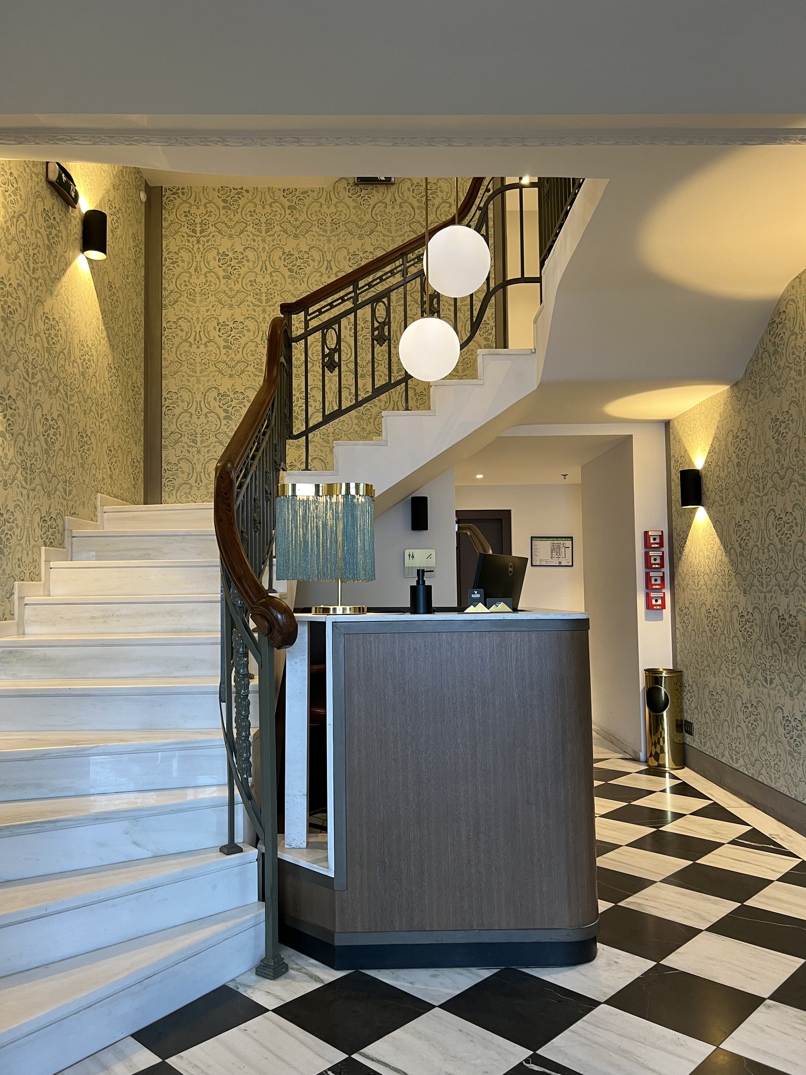 A staircase with a dark iron balustrade curving up past a wall, globe lights hanging in the stairwell