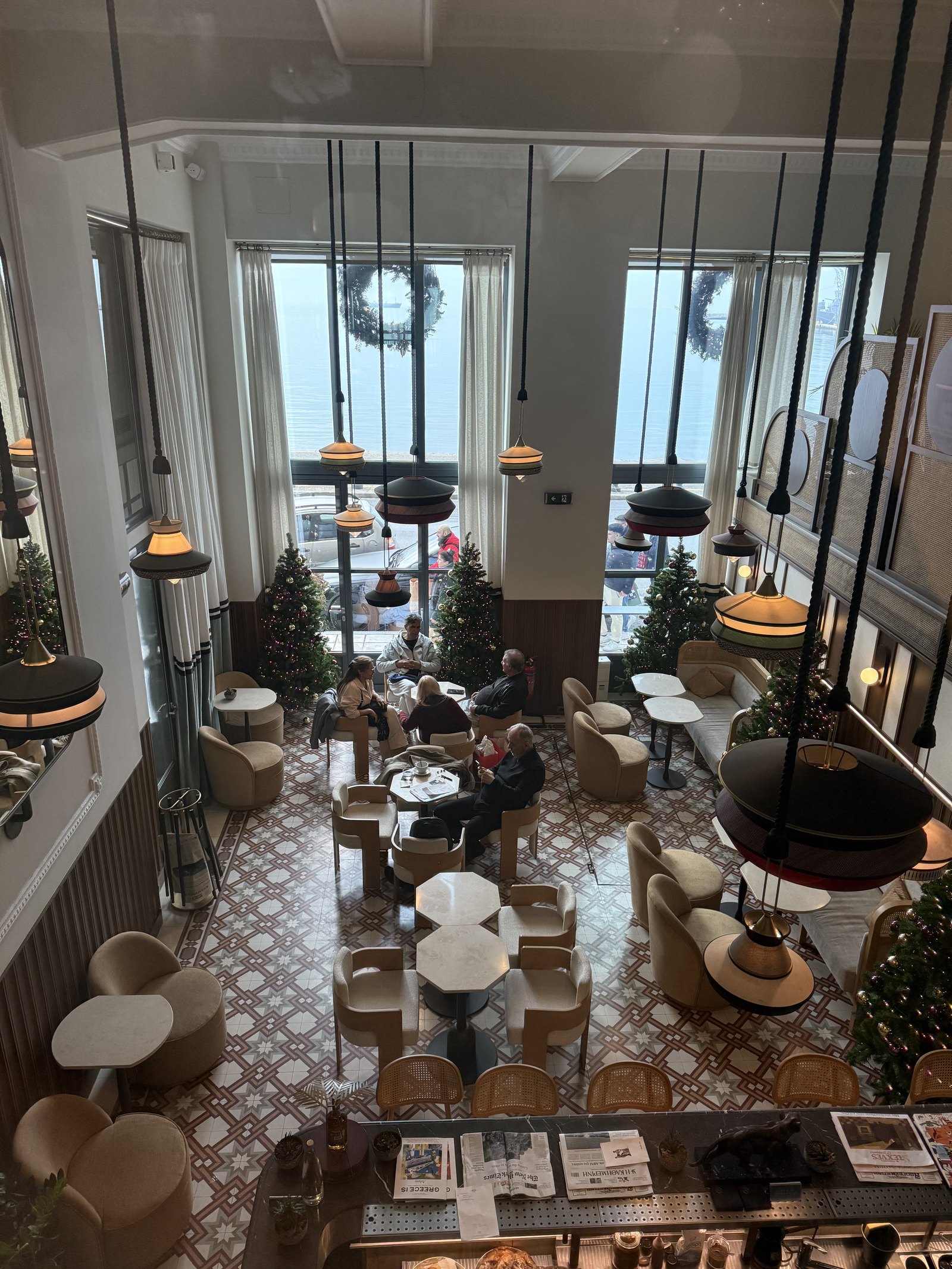 The double-height Tiger Loop cafe at ON Residence seen from the floor above, a patterned tile floor, hanging lamps, small Christmas trees and the Thermaic Gulf through tall windows