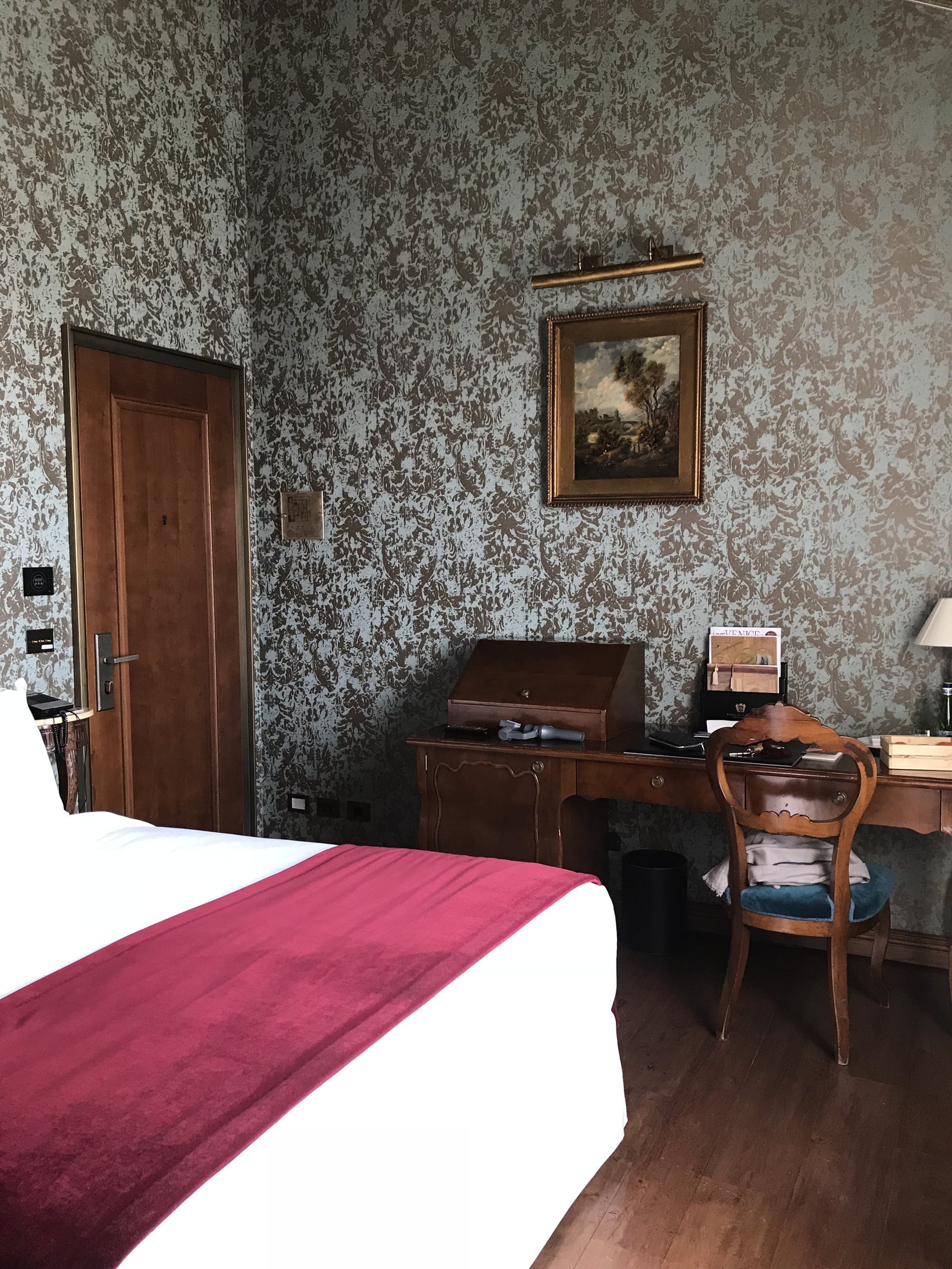 A bed with a red runner across white linen, a writing desk and chair beyond it against patterned wall silk