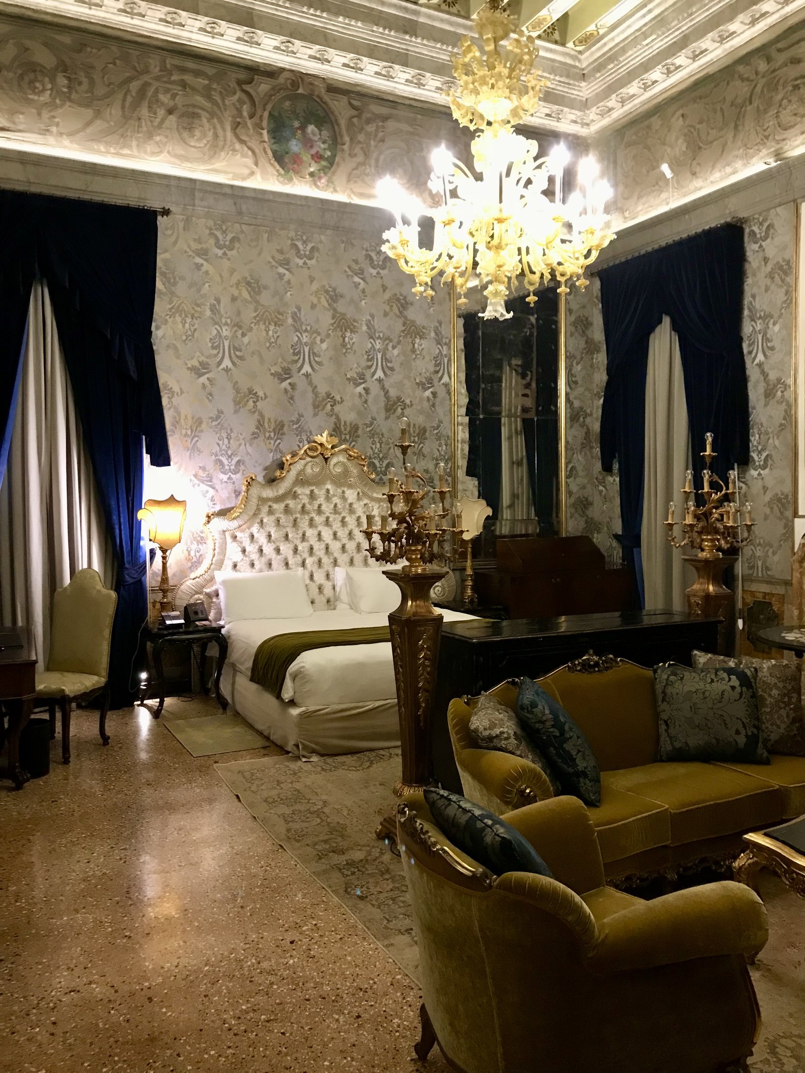 A room in silver and green damask with a Murano chandelier, a gilt bed with a tufted headboard and an olive velvet sofa