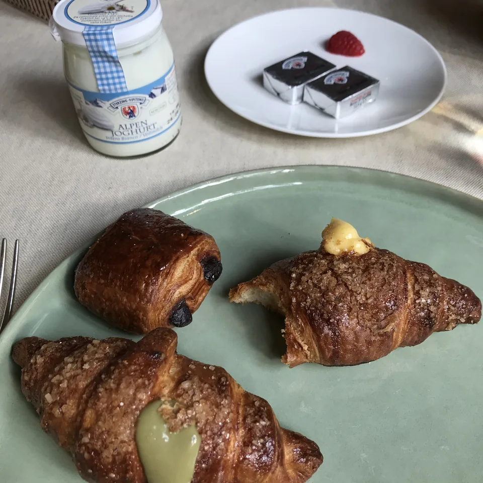 Two croissants on a pale green plate beside a jar of yoghurt and a small plate of chocolates with a raspberry