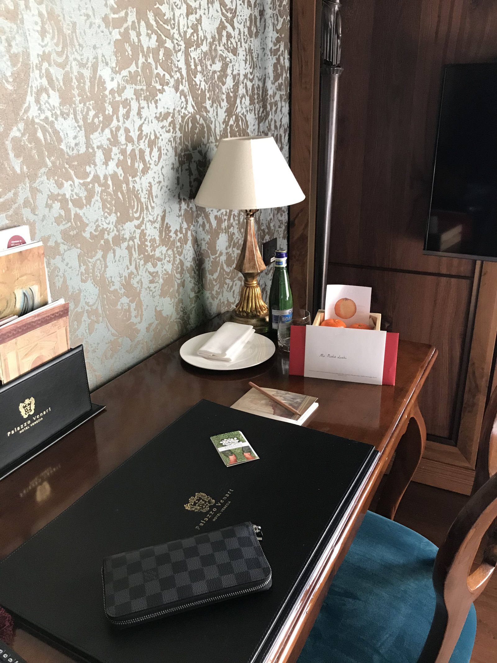 A desk with a brass lamp, a bottle of water, a welcome card and a leather folder on a dark top