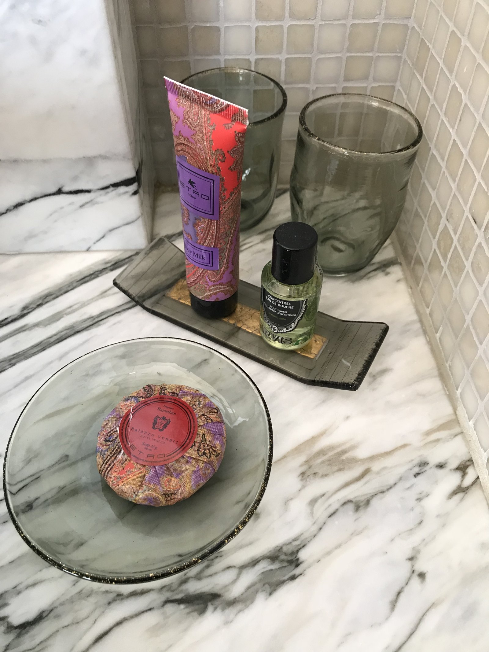 A tube of Etro bath product and a small bottle on a glass tray beside a wrapped soap on marble
