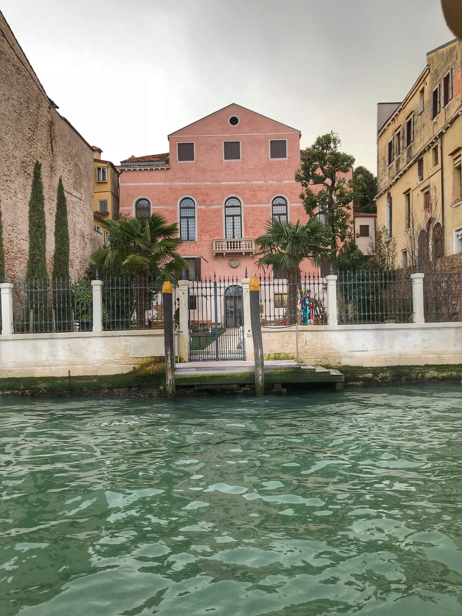 A pink palazzo seen from the Grand Canal behind its own garden fence, two palms and a wooden landing stage with mooring poles in front of it