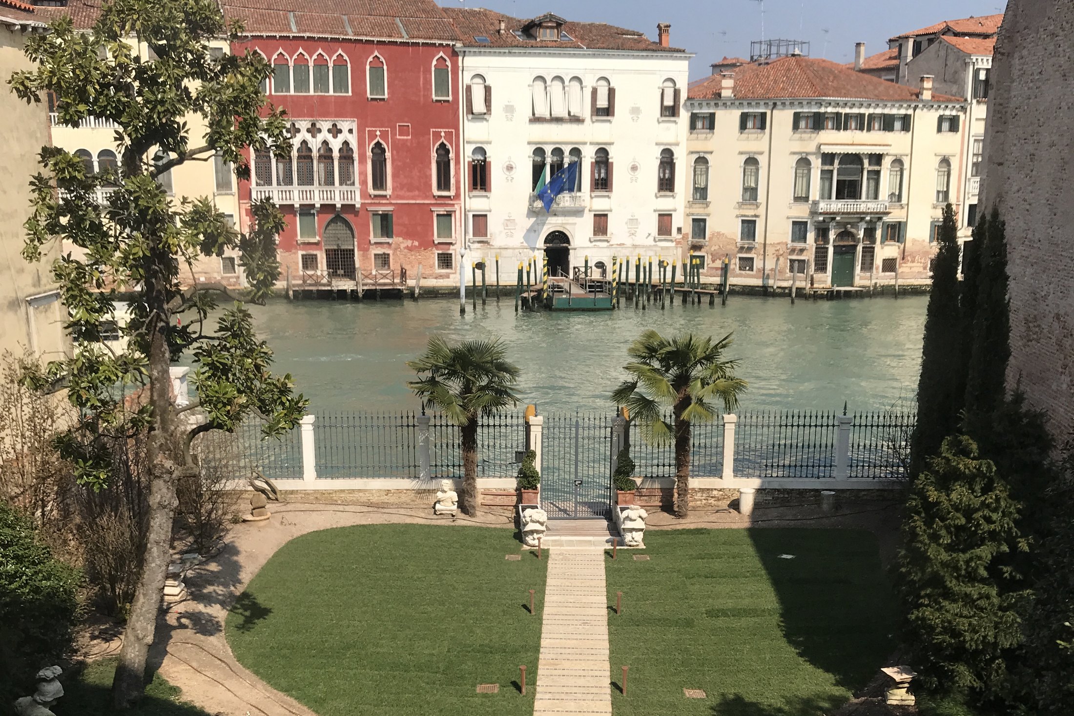 A lawn and a stone path running down between two palms to a gate on the Grand Canal, Gothic palazzi on the far bank