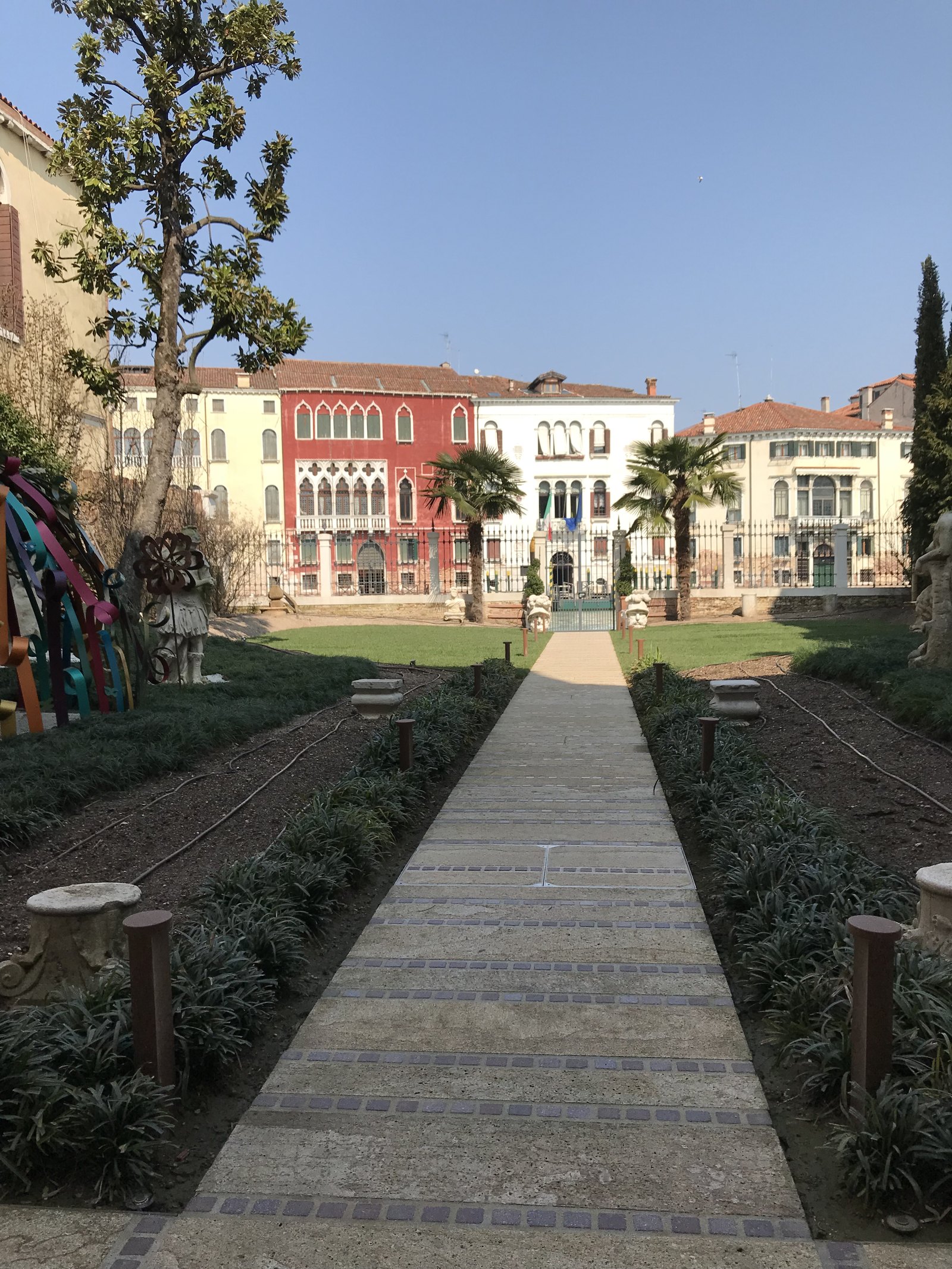 A stone path between lawns running towards a gate, with Gothic palazzi in red and white on the far side of the canal