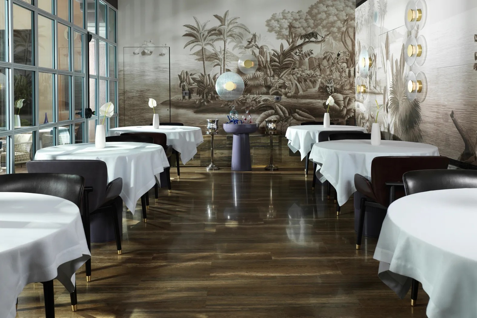 A dining room with laid tables in white linen, a tall window wall onto a courtyard along the left and a wall painted in sepia with palms, a leopard, a tiger and a lion, glass sconces down the right