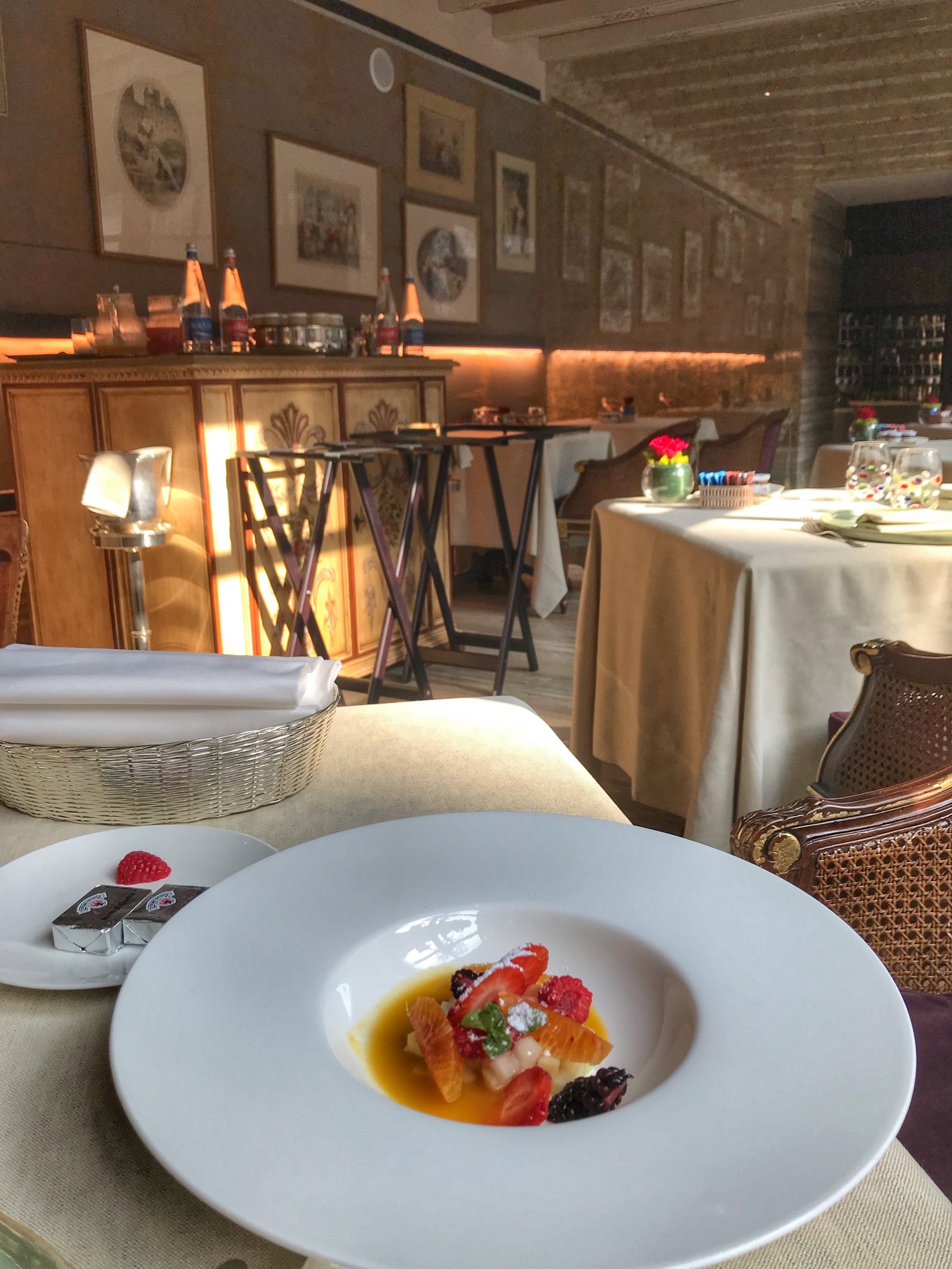 A dining room with laid tables in cream linen, framed prints on a brick wall, service trolleys in the corner and a plate of fruit in the foreground