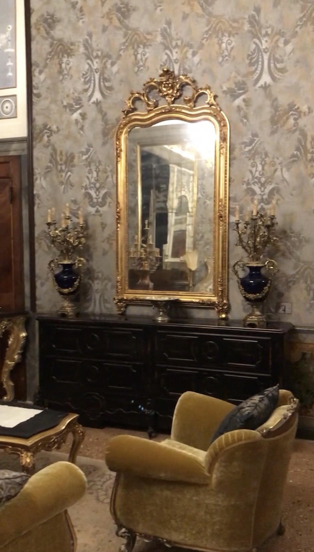 A gold framed mirror and candelabra above a dark carved commode, with yellow velvet armchairs on a patterned carpet