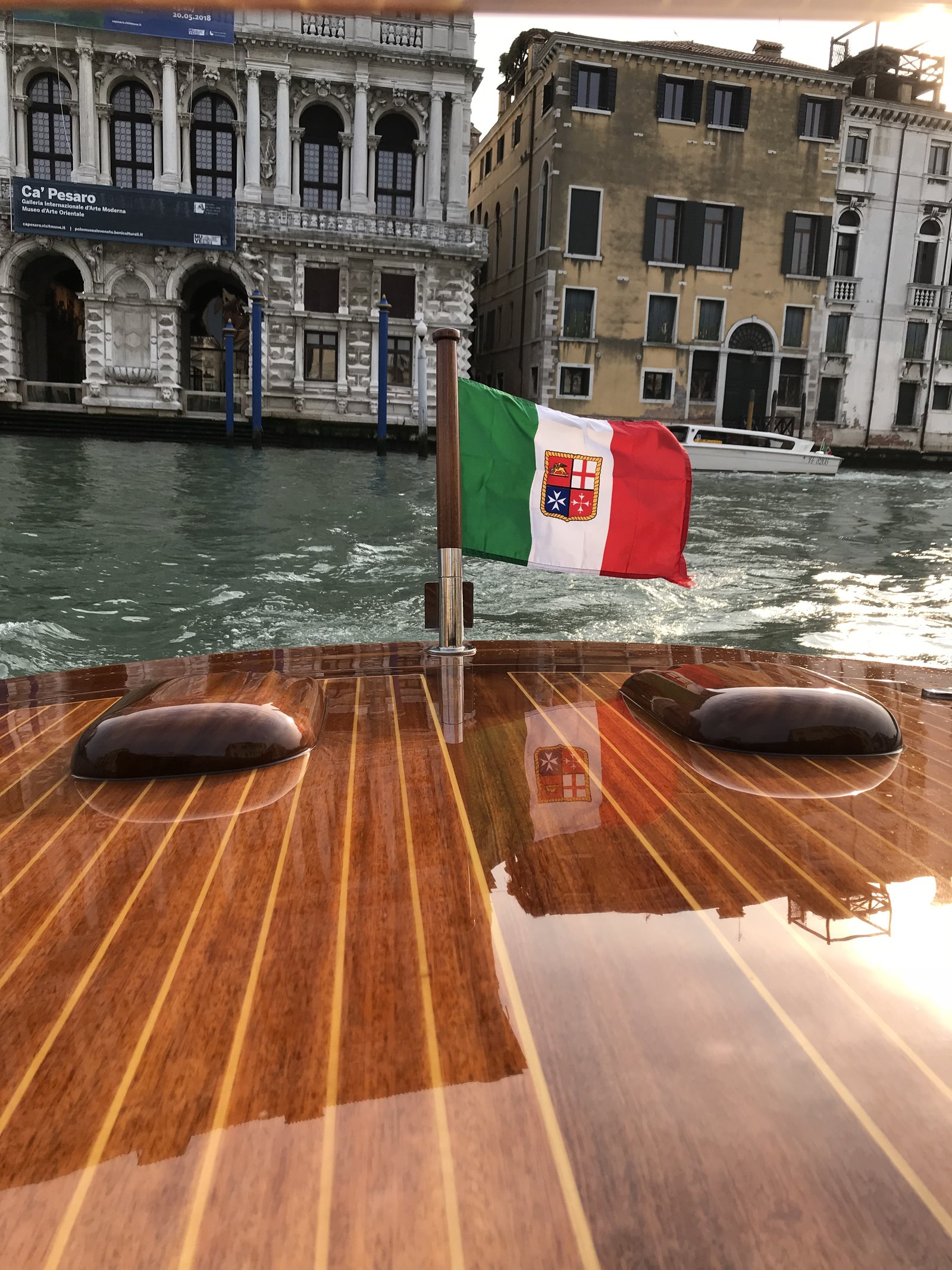 The varnished deck of a water taxi with an Italian flag at the stern, Ca' Pesaro on the far bank of the Grand Canal