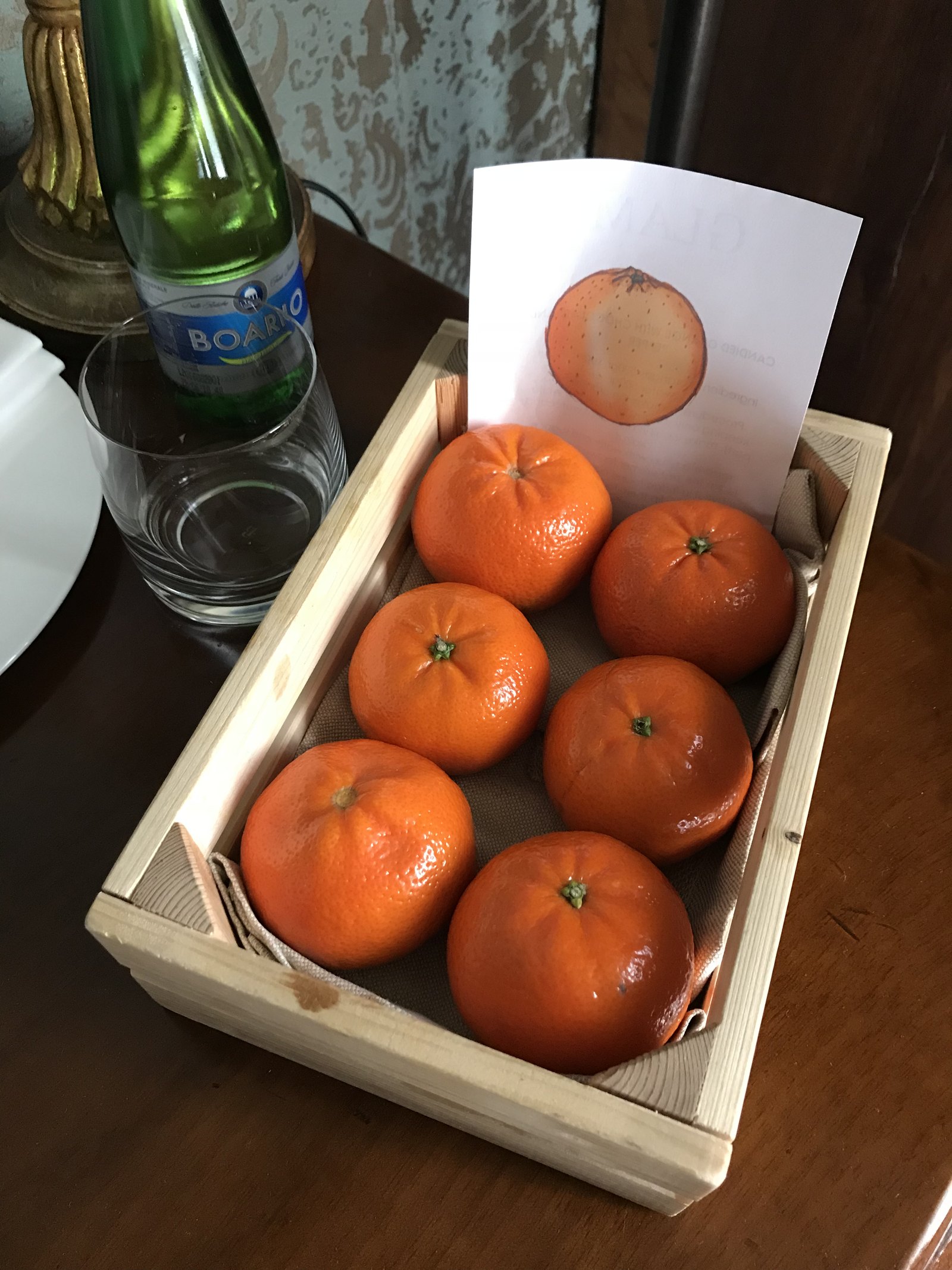 A wooden crate of six oranges with a printed card beside a bottle of water and two glasses