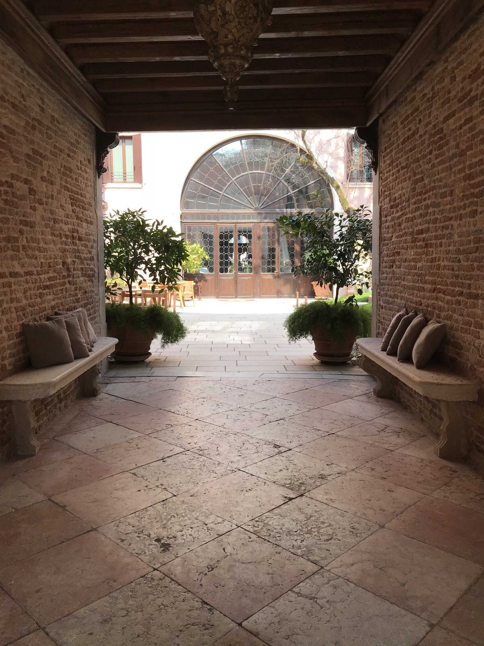 A brick passage with a beamed ceiling and stone benches opening onto a glazed garden room