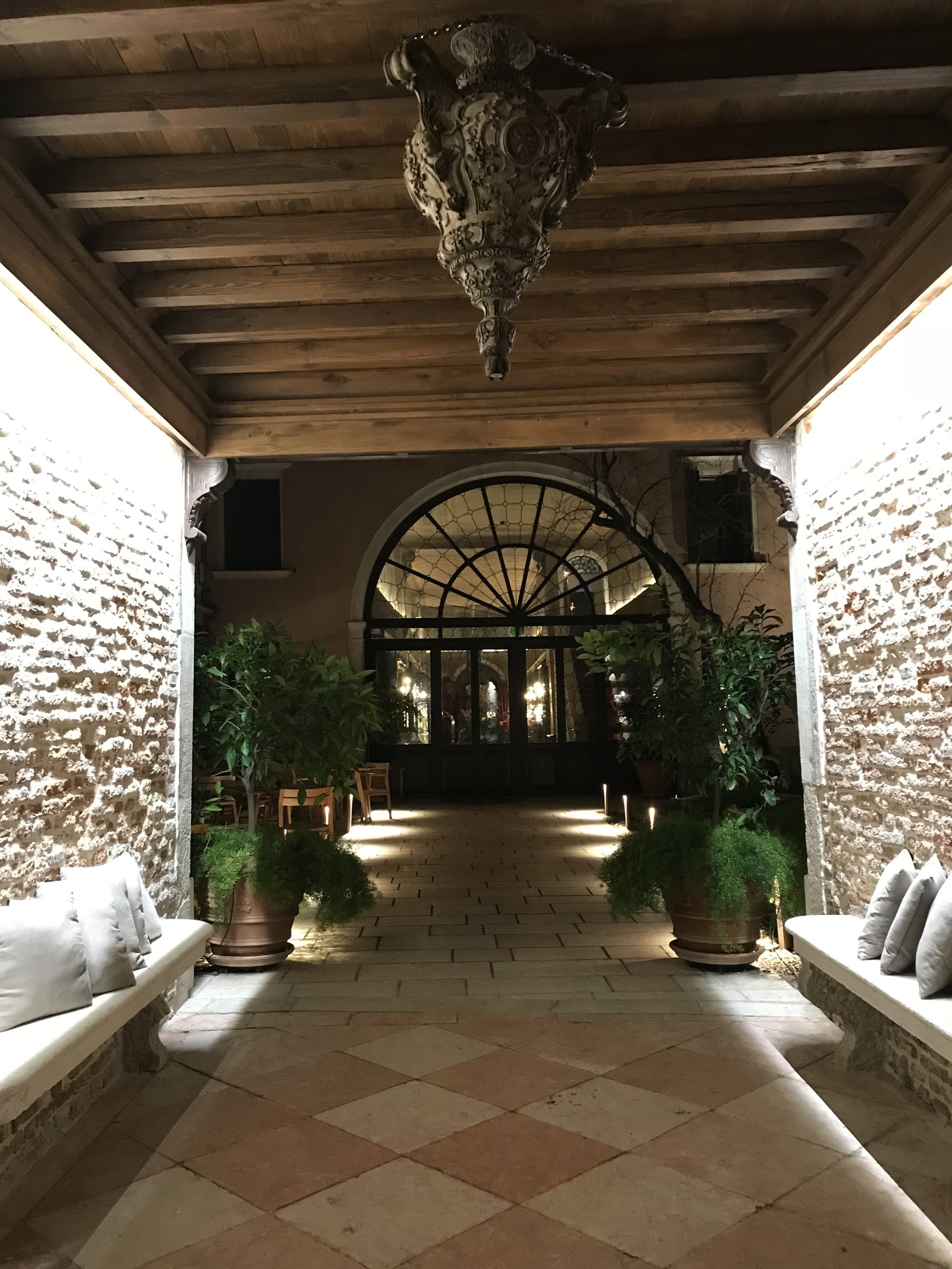 A brick passage lit at night, with cushioned stone benches, potted plants and a great arched glass door glowing at the far end