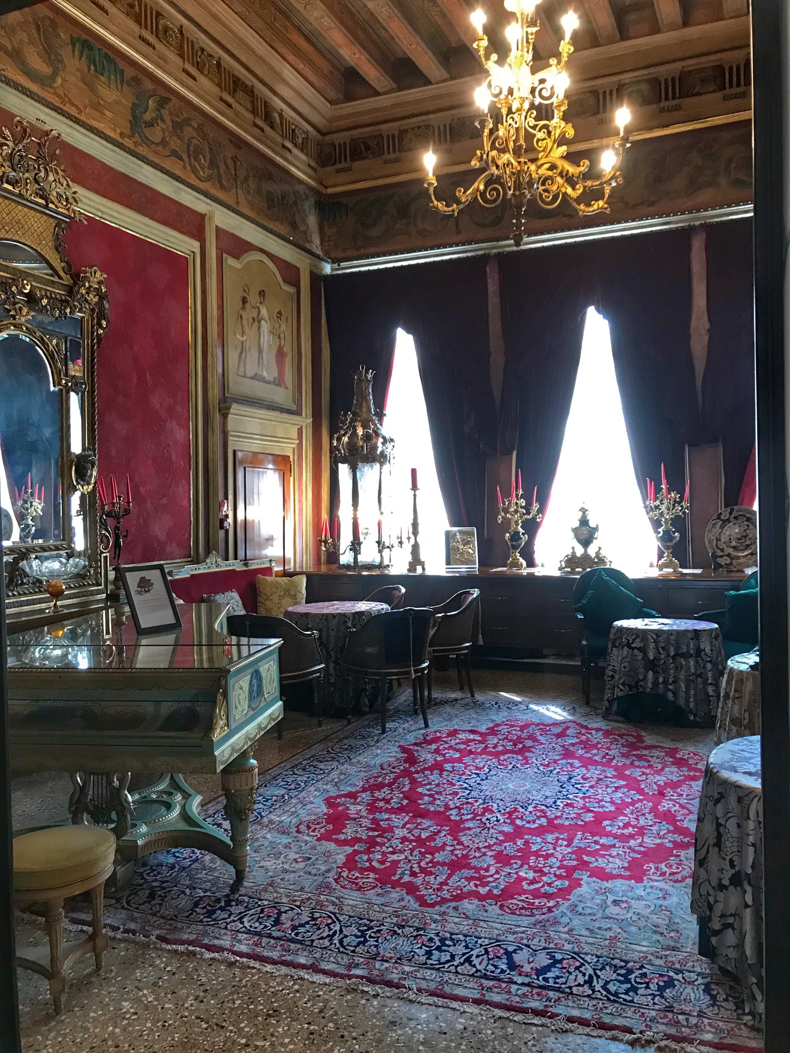 A salon with red damask walls, a Murano chandelier, gilt mirrors and a deep pink carpet between tall curtained windows