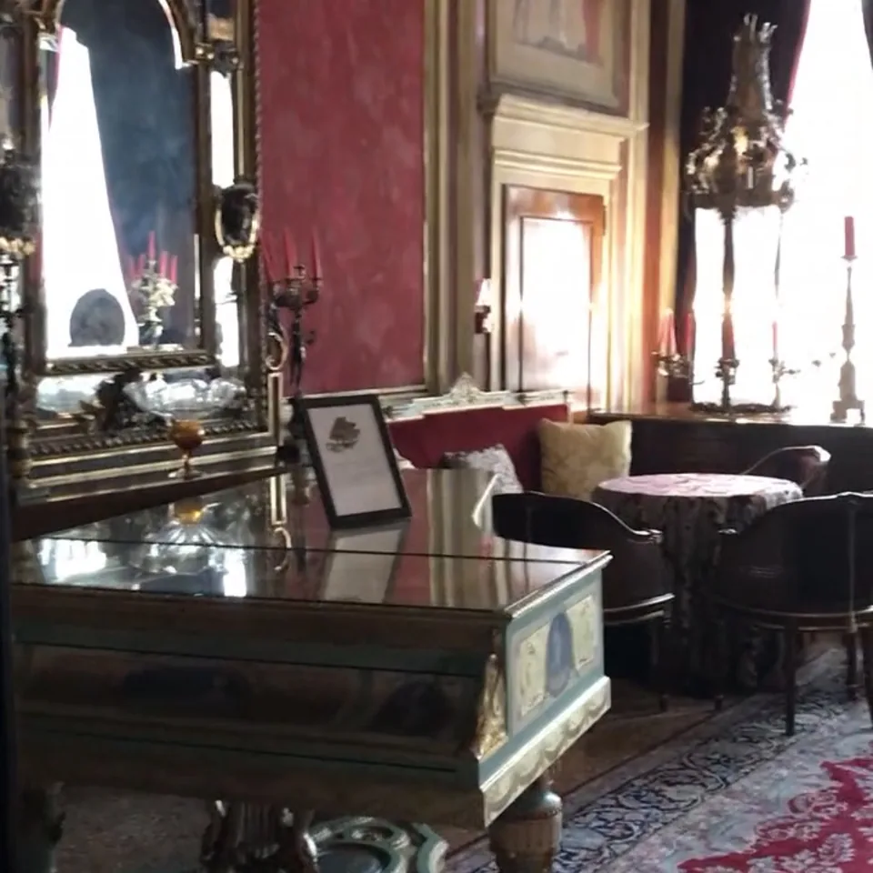 A salon in red damask in the evening, a gilt mirror over a mirrored table with candlesticks and a painted overdoor