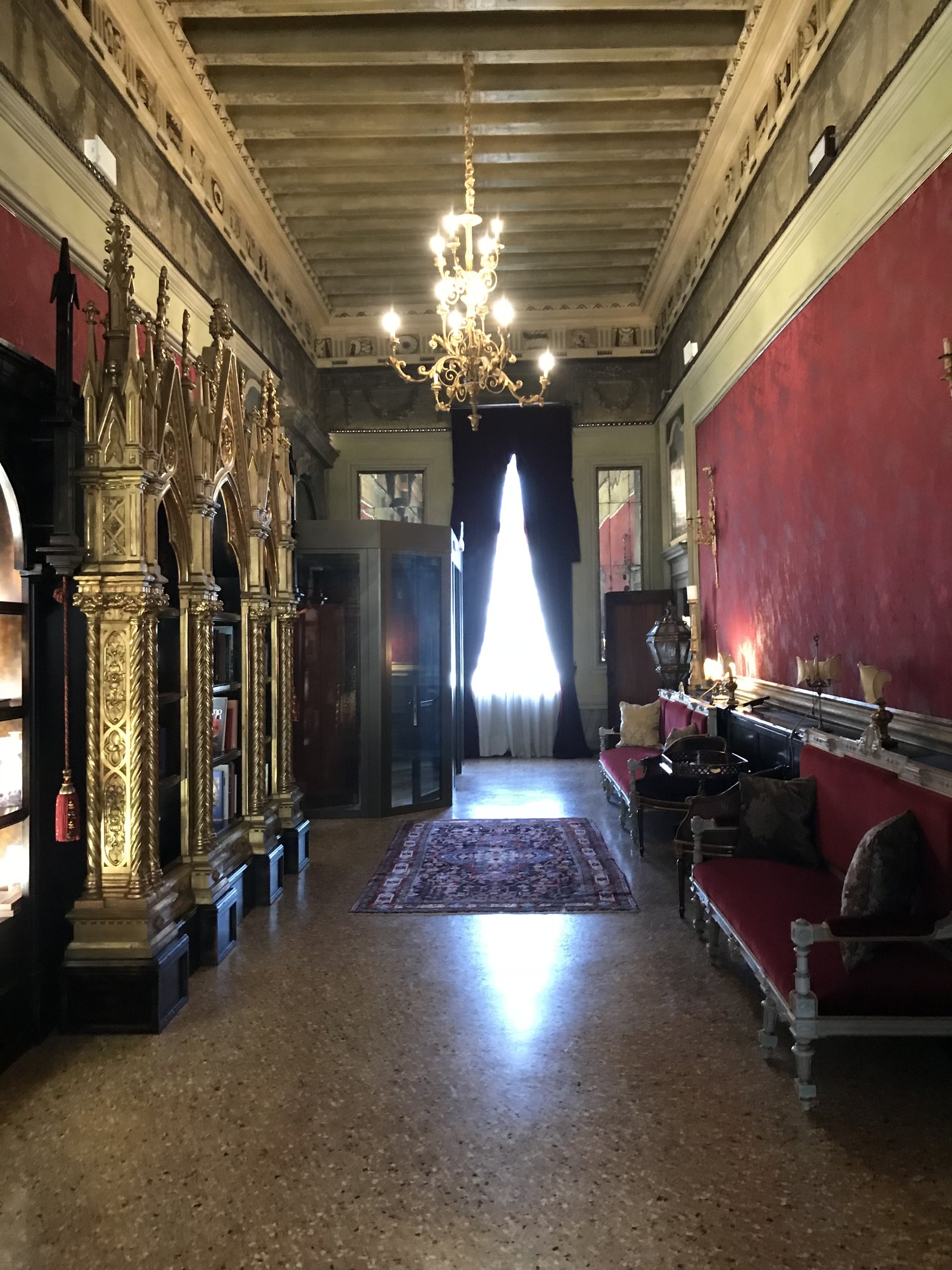 A long room in red damask with a chandelier down its length, a bench along one wall and a lit window at the far end