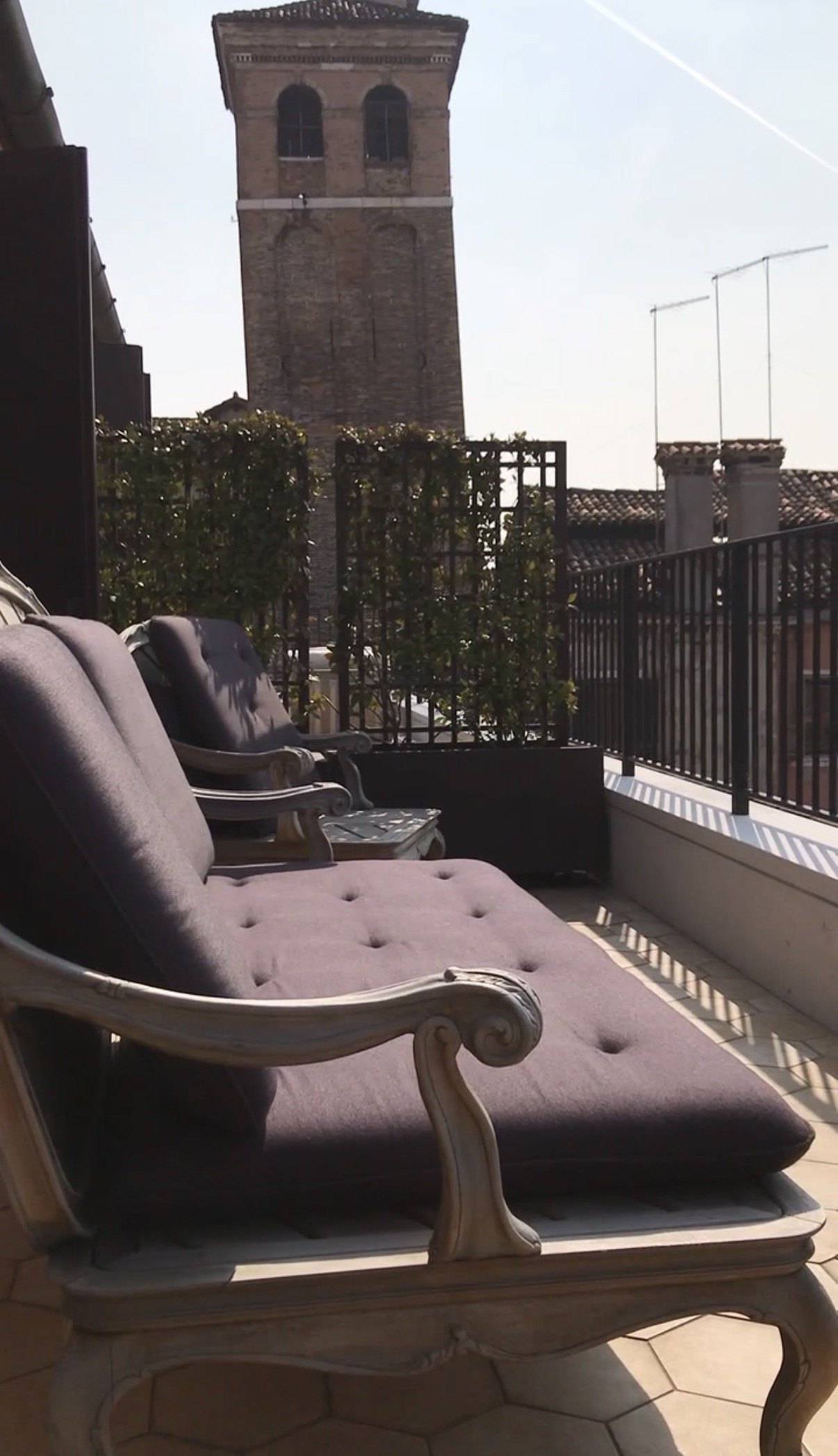 Two long painted chairs with deep cushions on a tiled terrace, a brick campanile rising behind a trellis