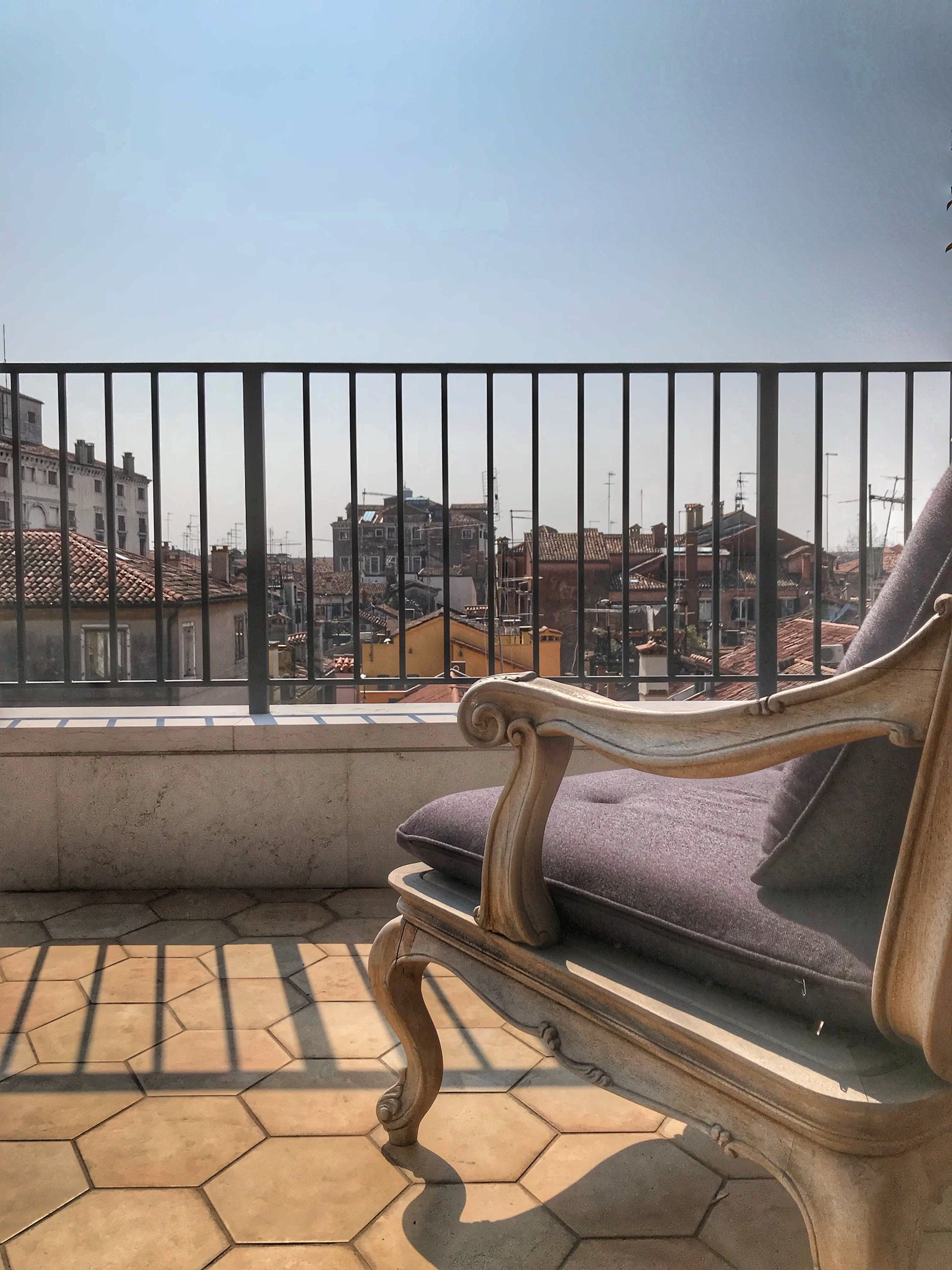 A painted chaise longue on hexagonal terracotta tiles behind an iron railing, the tiled roofs of Venice beyond