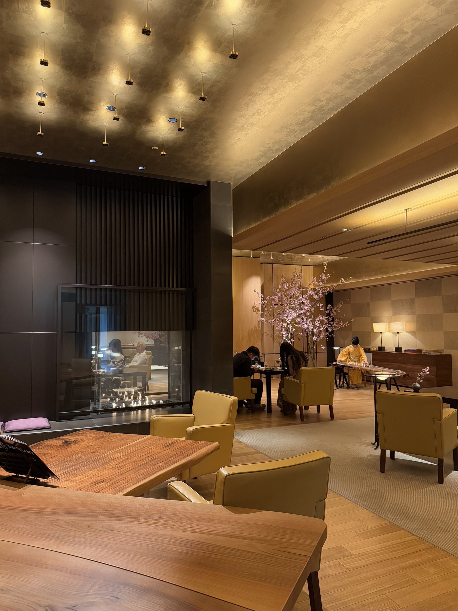A lounge with yellow armchairs around a low table and a fire in a stone wall