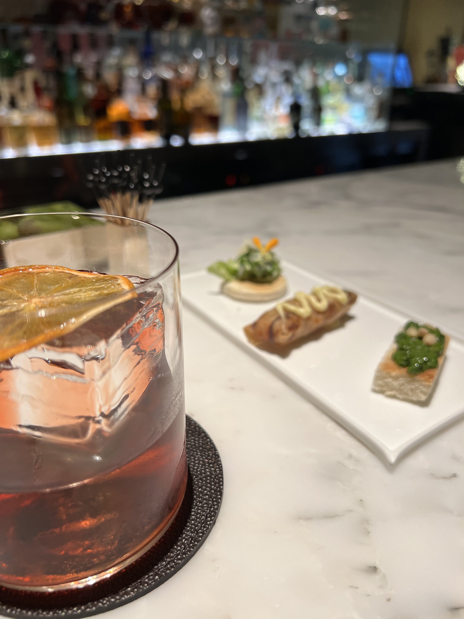 A cocktail with an orange twist in a rocks glass on a marble table, beside a plate of canapés