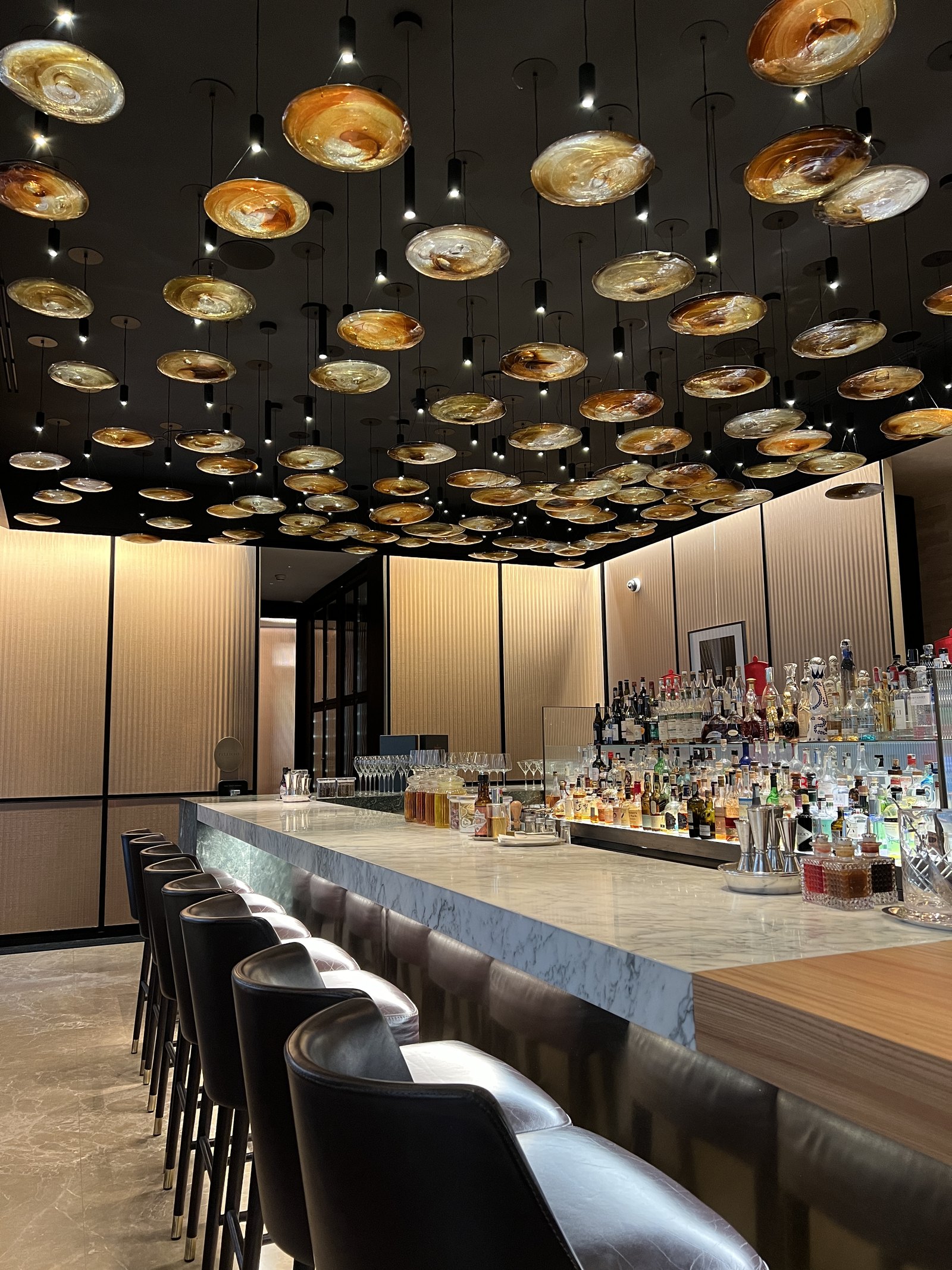 A marble bar with rows of bottles behind it and a ceiling of golden pendant lamps