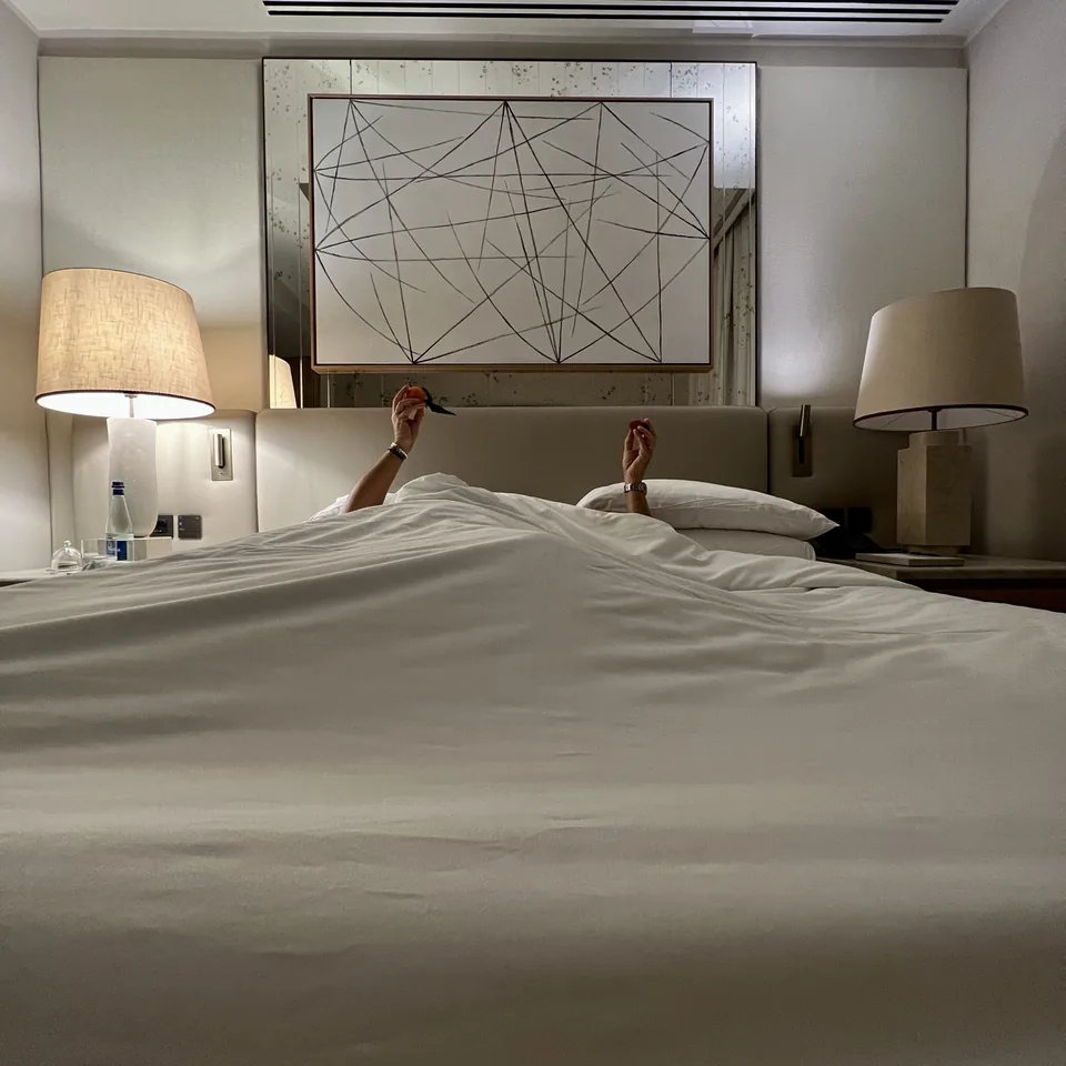 A made bed with white linen and two lamps, seen from the foot, with two arms raised from under the covers holding clementines