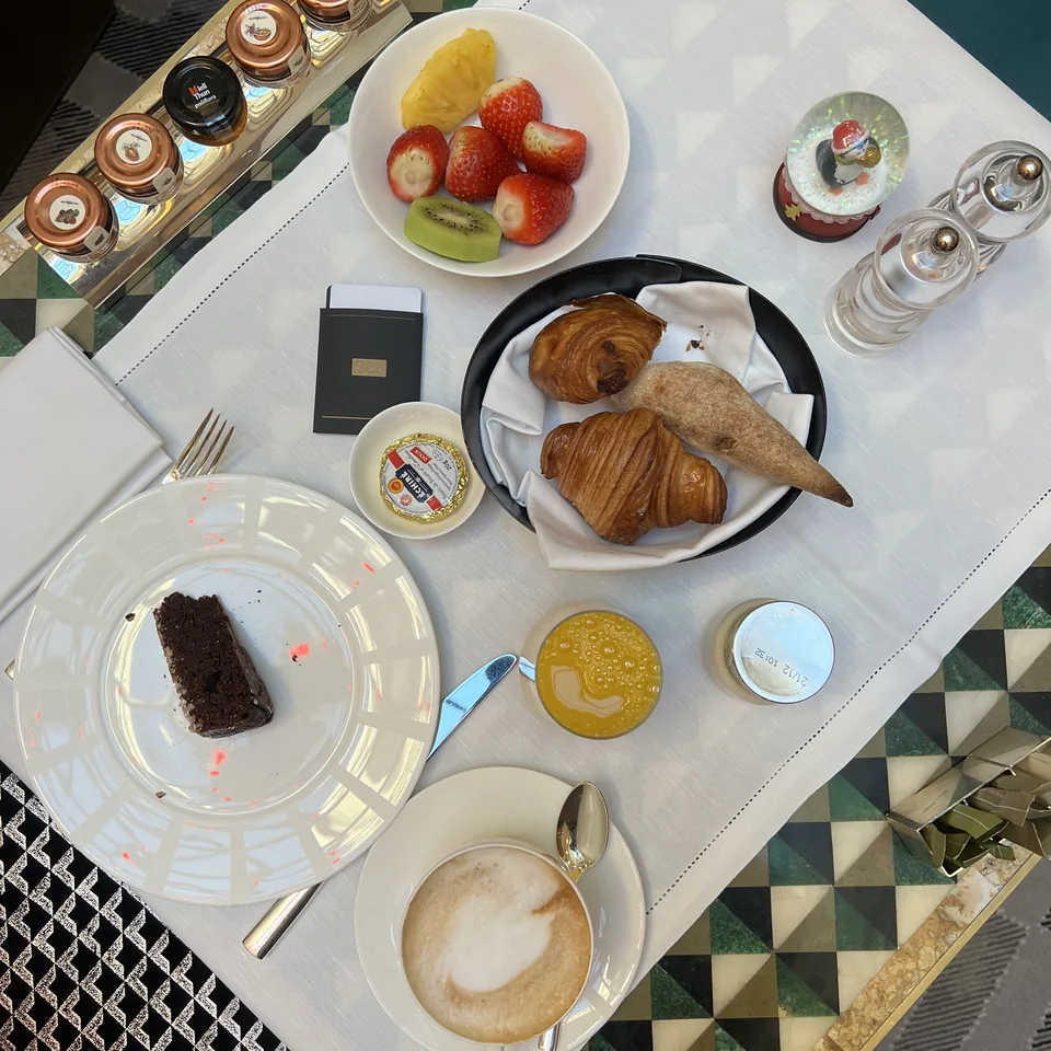 Breakfast on a patterned table seen from above: croissants, a cake, a cup of coffee and fruit