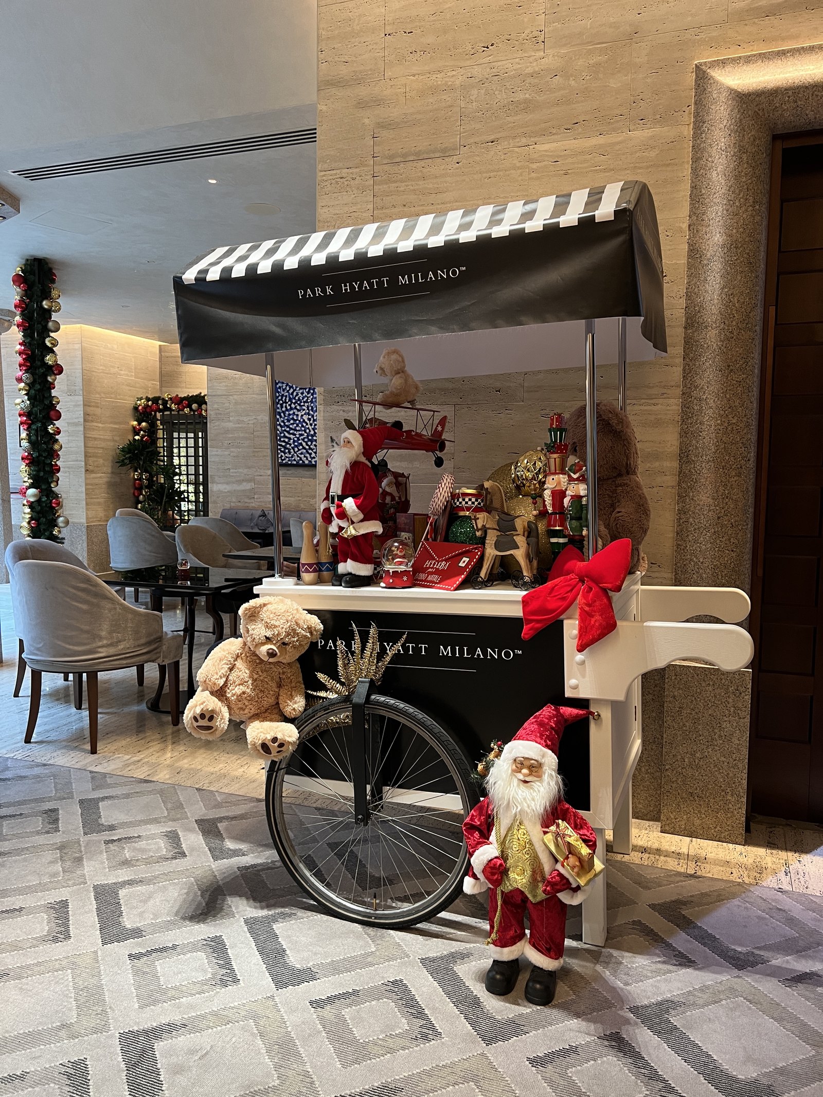A Santa Claus and a teddy bear on a red cart in the lobby, beside a stair lined with garland