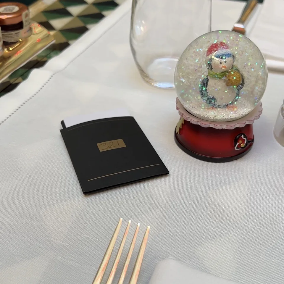 A table laid for dinner with a clear glass, a snow globe with a snowman on a red base, and a black place card marked 321
