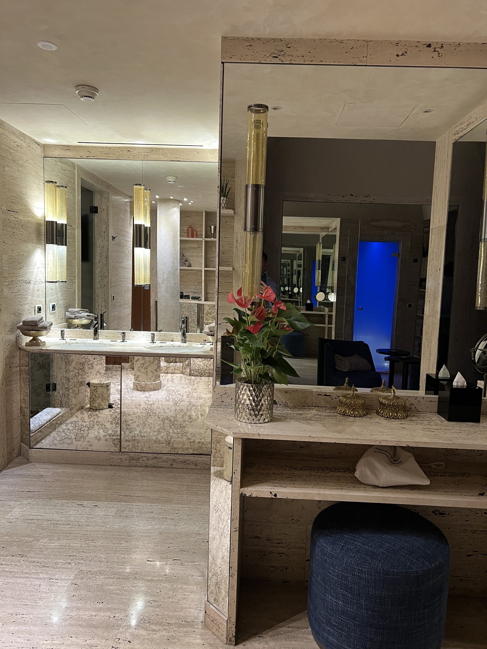 A marble washroom with a vanity, a dark stool and a mirror reflecting a blue door
