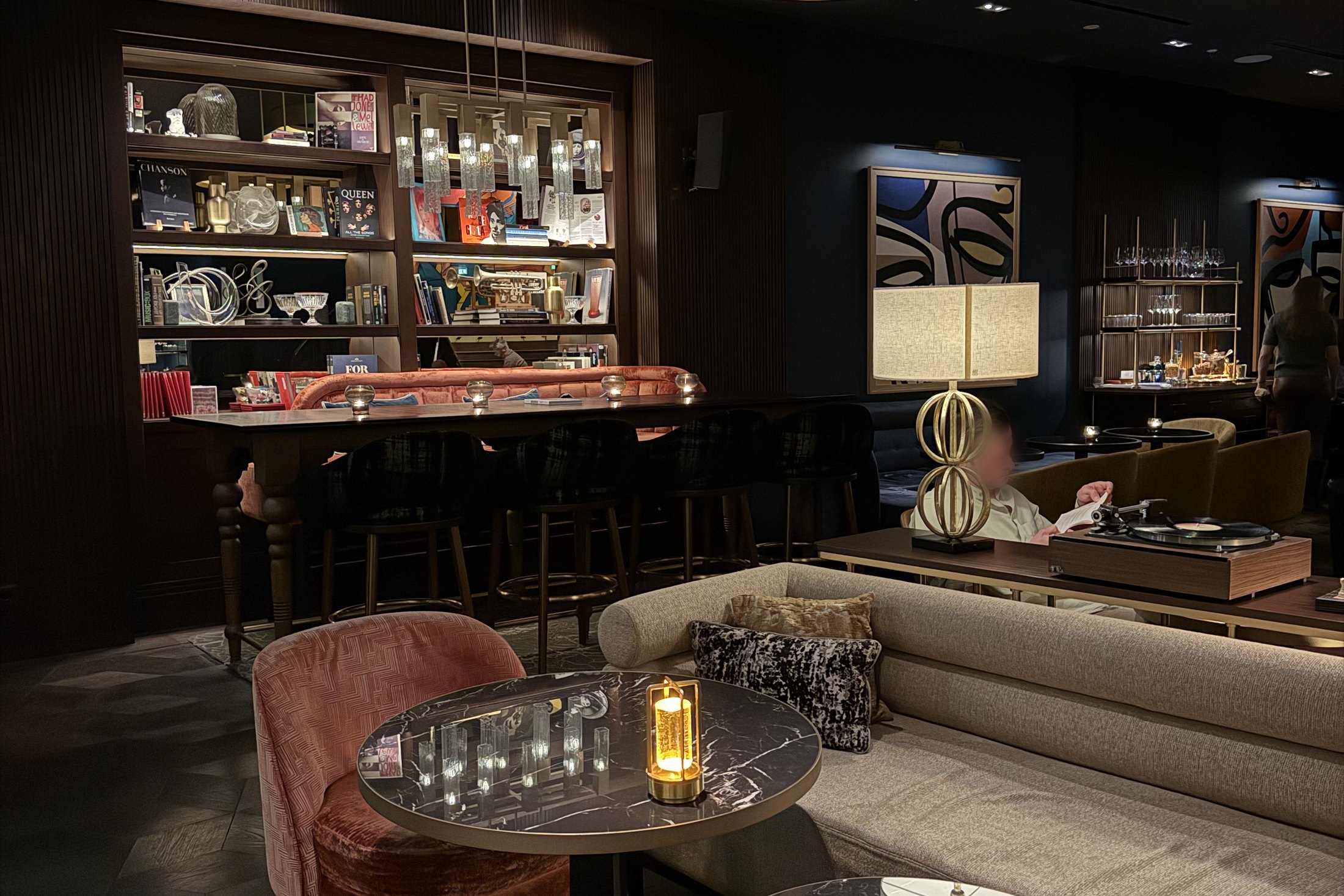 A dark bar room with velvet seating, round tables, a lit bottle display and low golden light