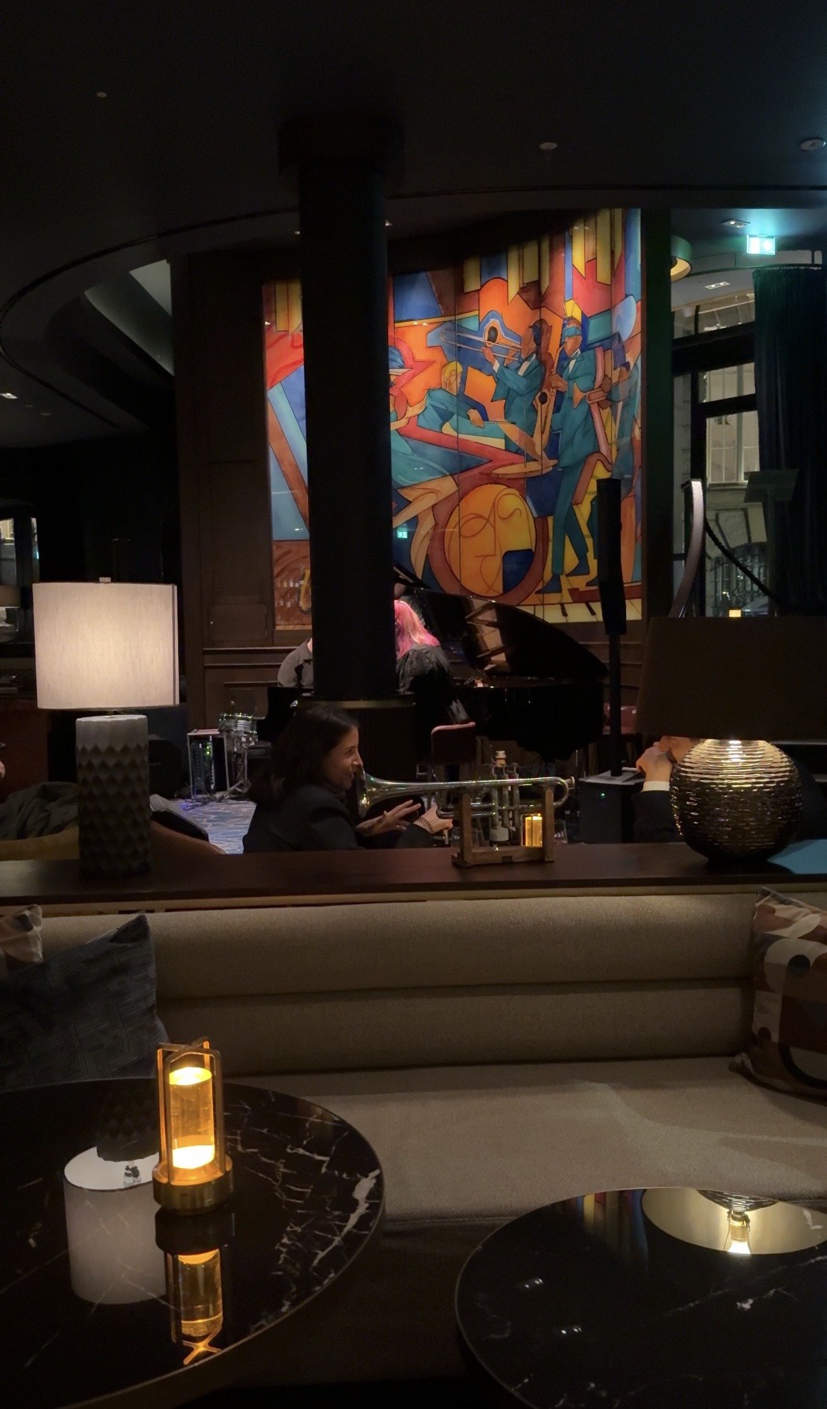 A bar with a large mural of musicians in bright cubist colour, a grand piano and banquettes below it