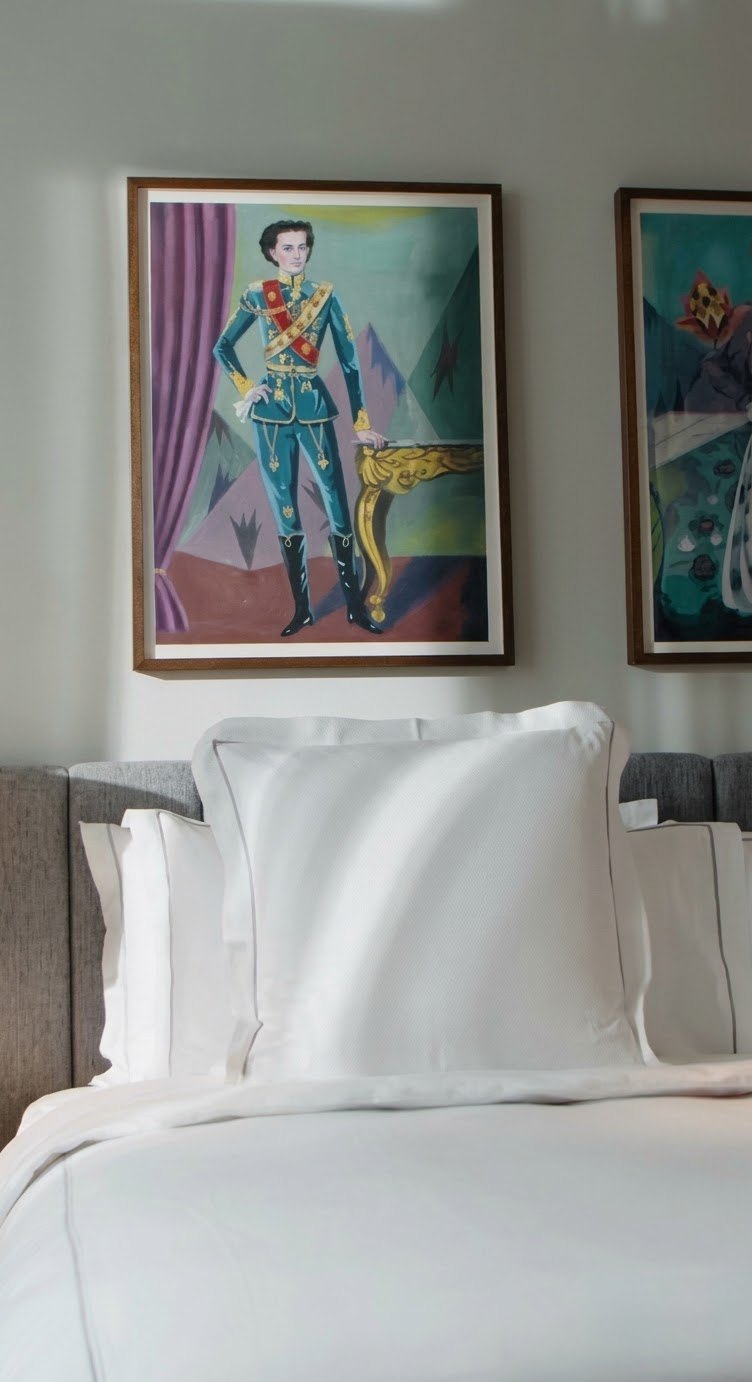 A bed with white pillows under a framed painting of a figure in Bavarian dress in flat graphic colour