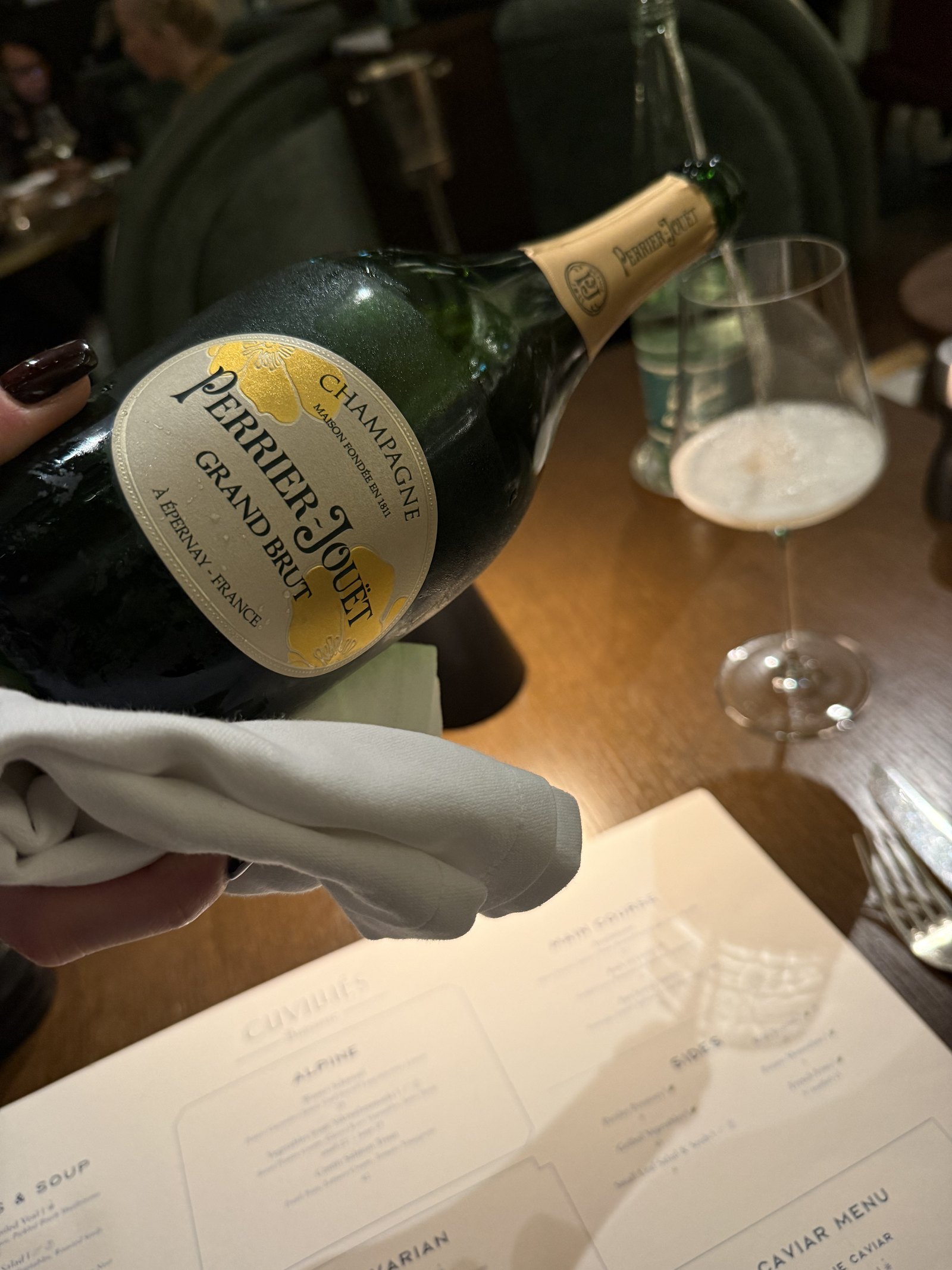 A bottle of Perrier-Jouët being poured into a glass beside a menu on a dark table