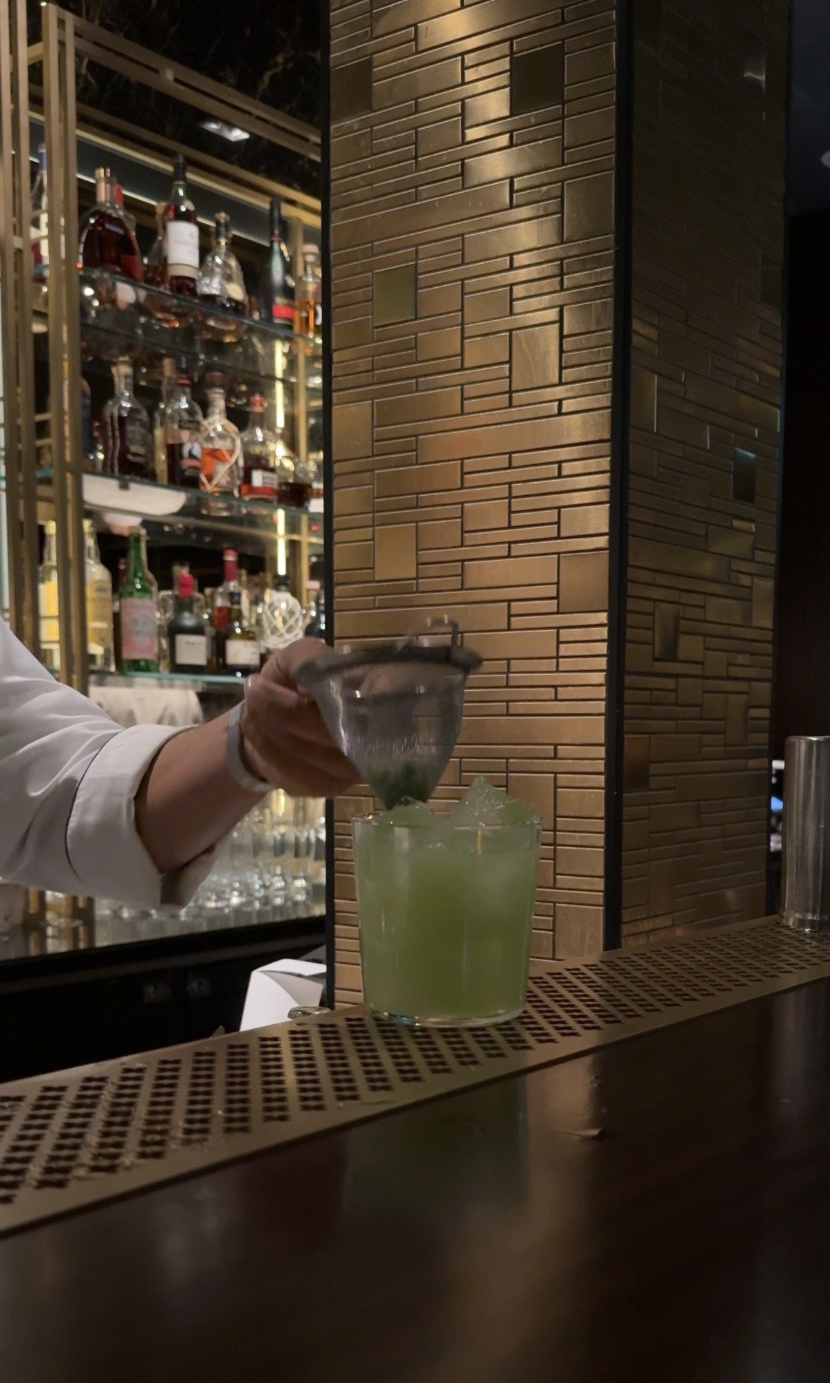 A pale green cocktail being set down on a dark counter by a hand in a white sleeve