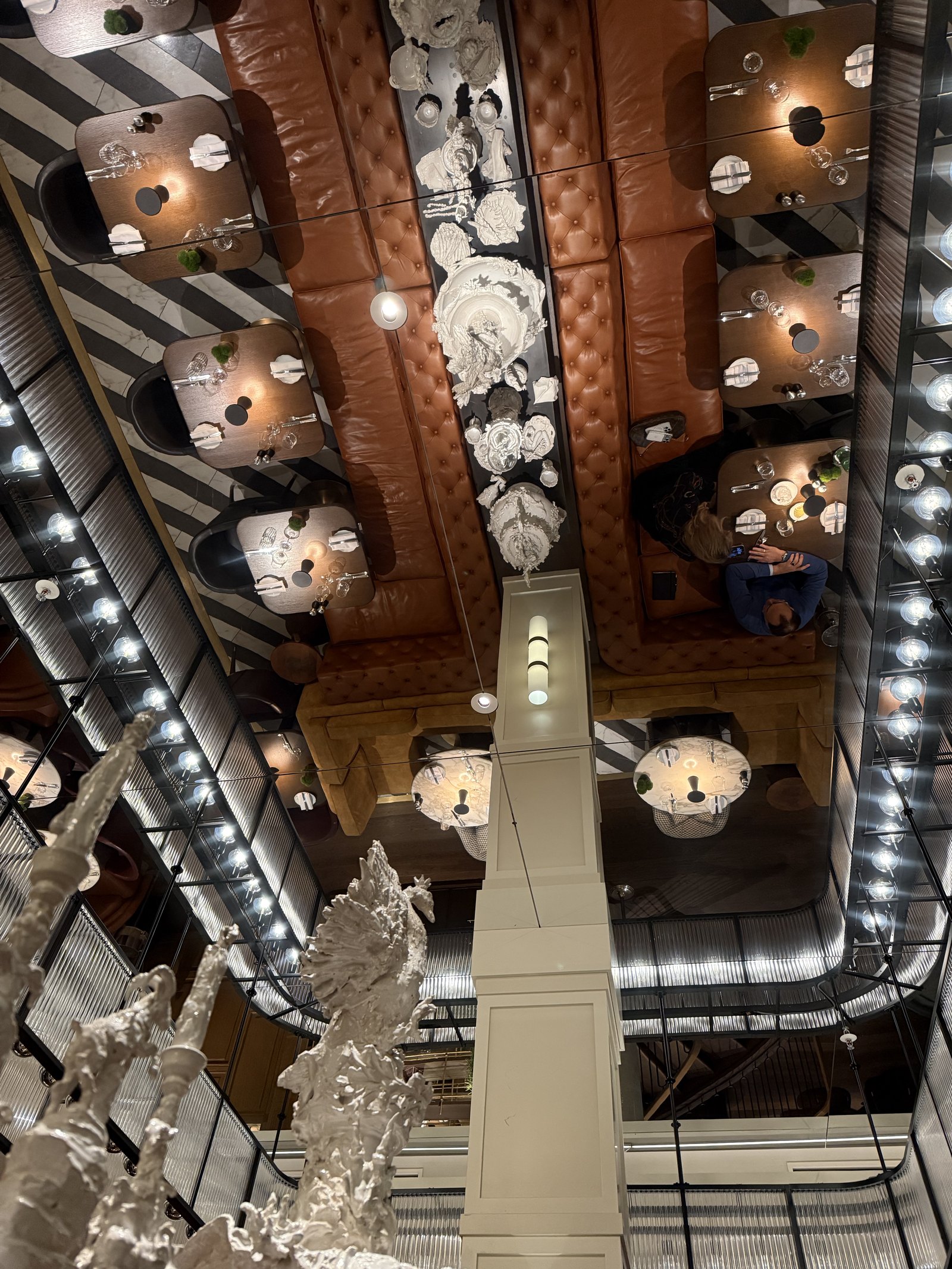 A mirrored ceiling reflecting laid tables, tan tufted banquettes and a line of white porcelain pieces, a white sculpture rising from below