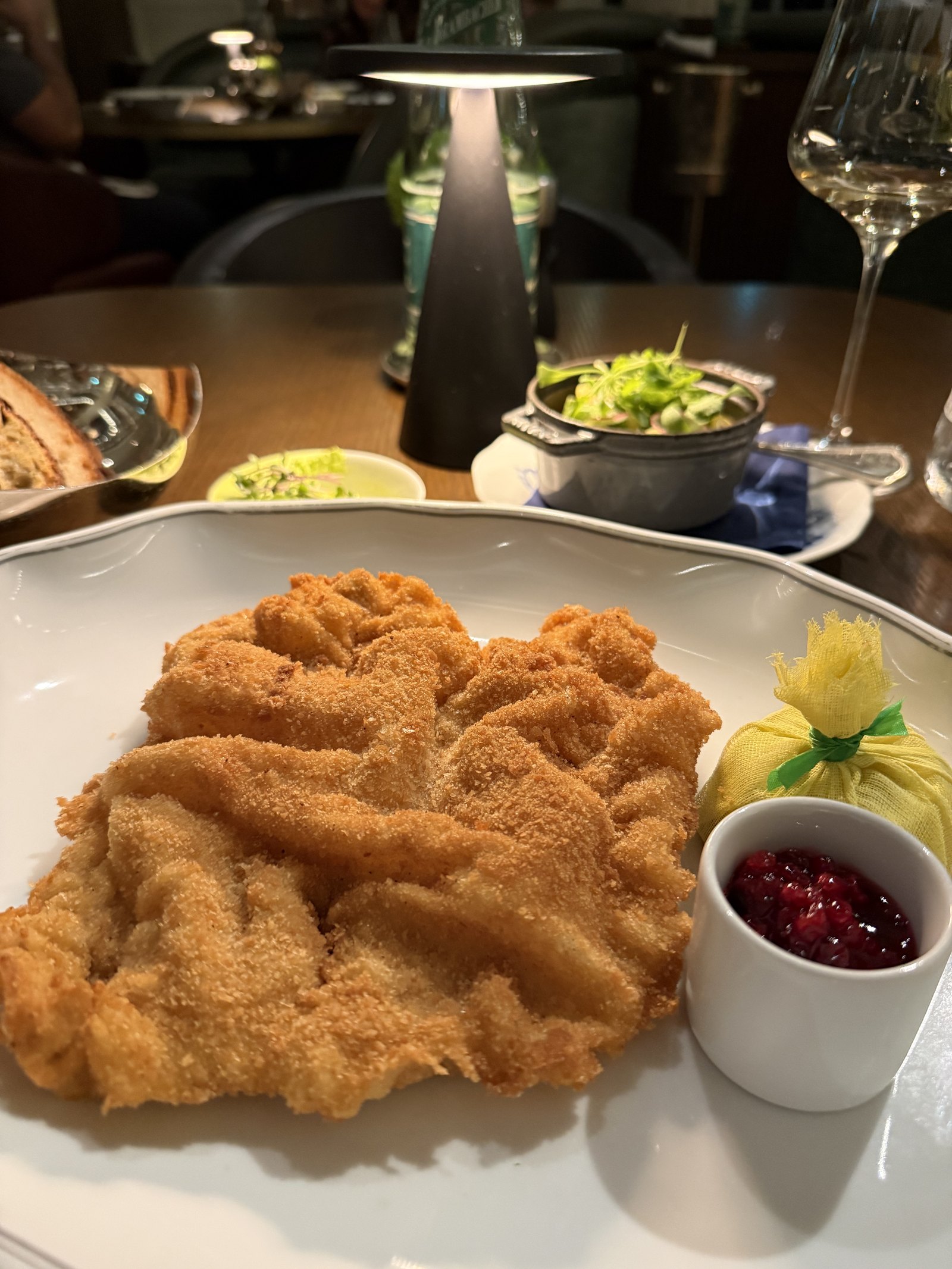 A large breaded veal schnitzel on a white plate with a dish of cranberry sauce and a lemon in paper