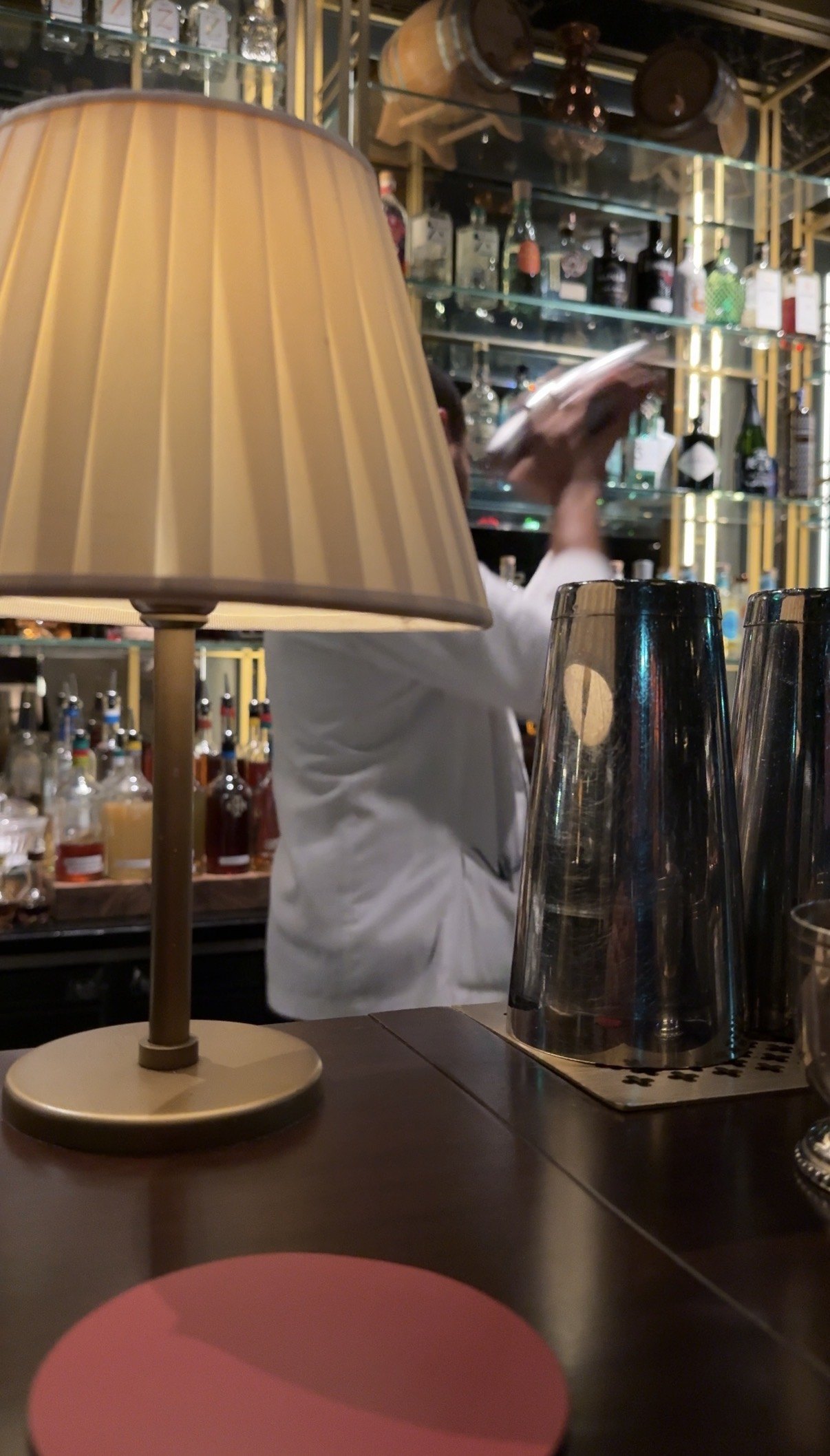 A shaker and a mixing glass on a counter under a pleated lampshade, bottles behind