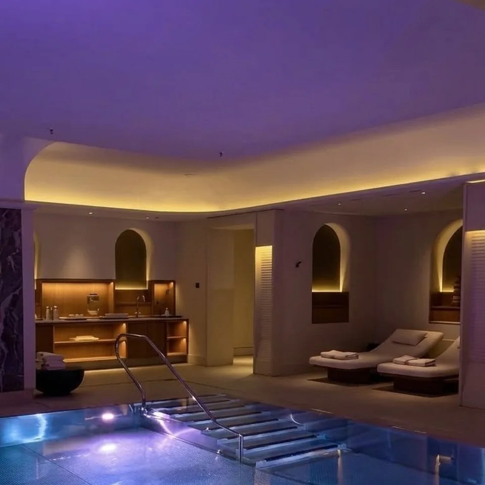 An indoor pool lit blue under a ceiling washed in violet light, loungers in arched alcoves along one side