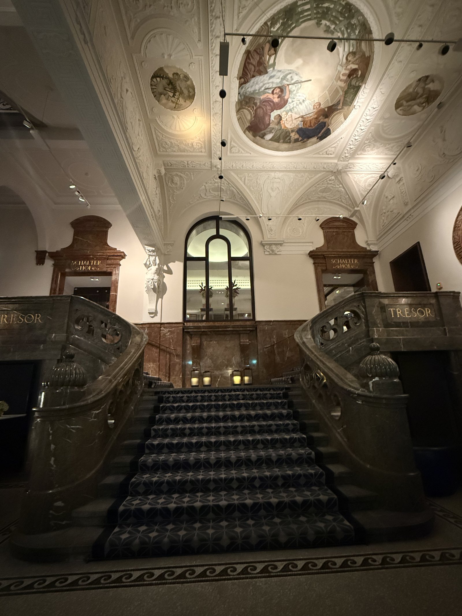 A banking hall staircase under a painted ceiling, TRESOR carved into the marble balustrades on both sides