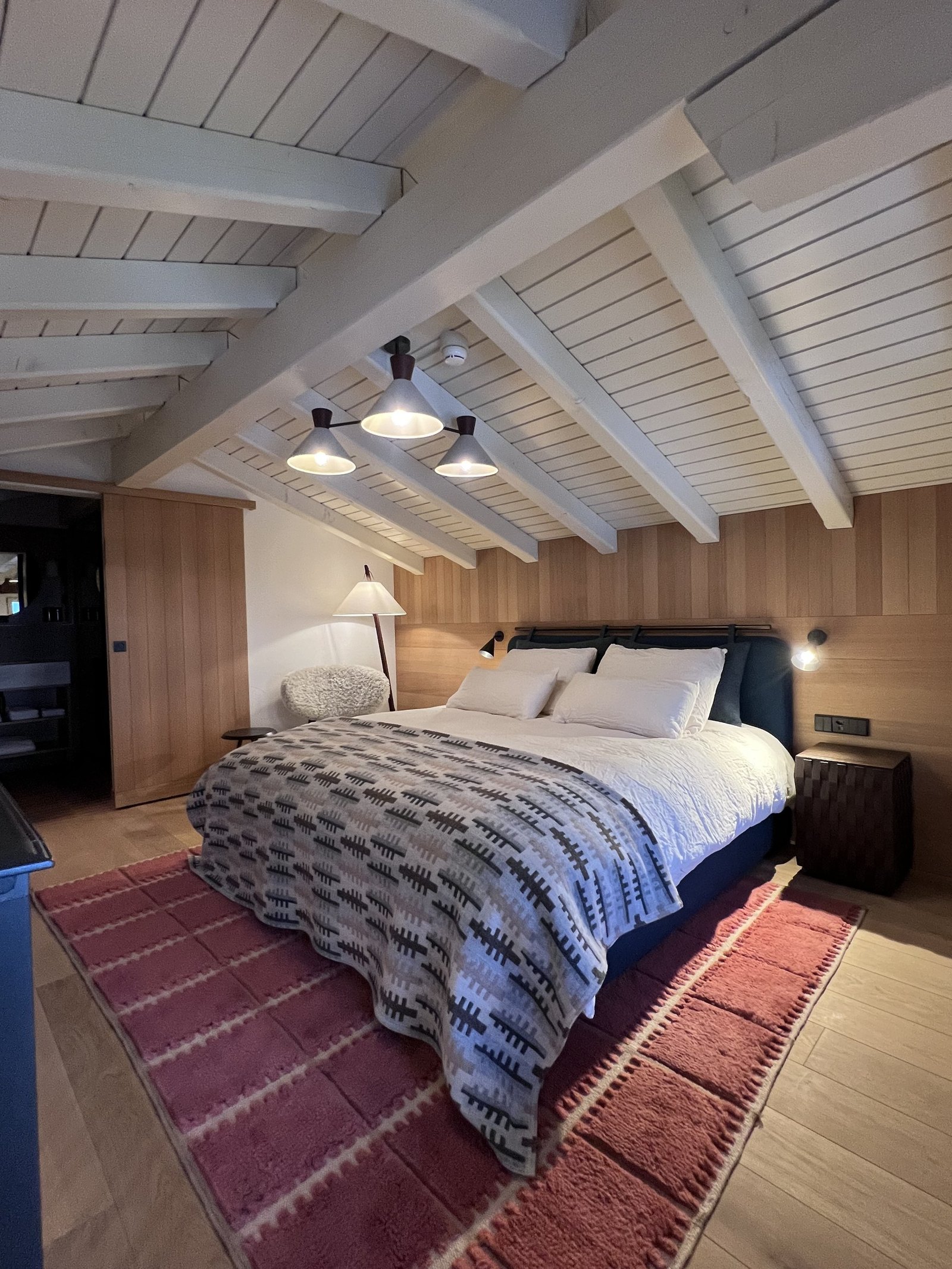 A bed under a white painted sloped ceiling with exposed beams and a grey knitted throw