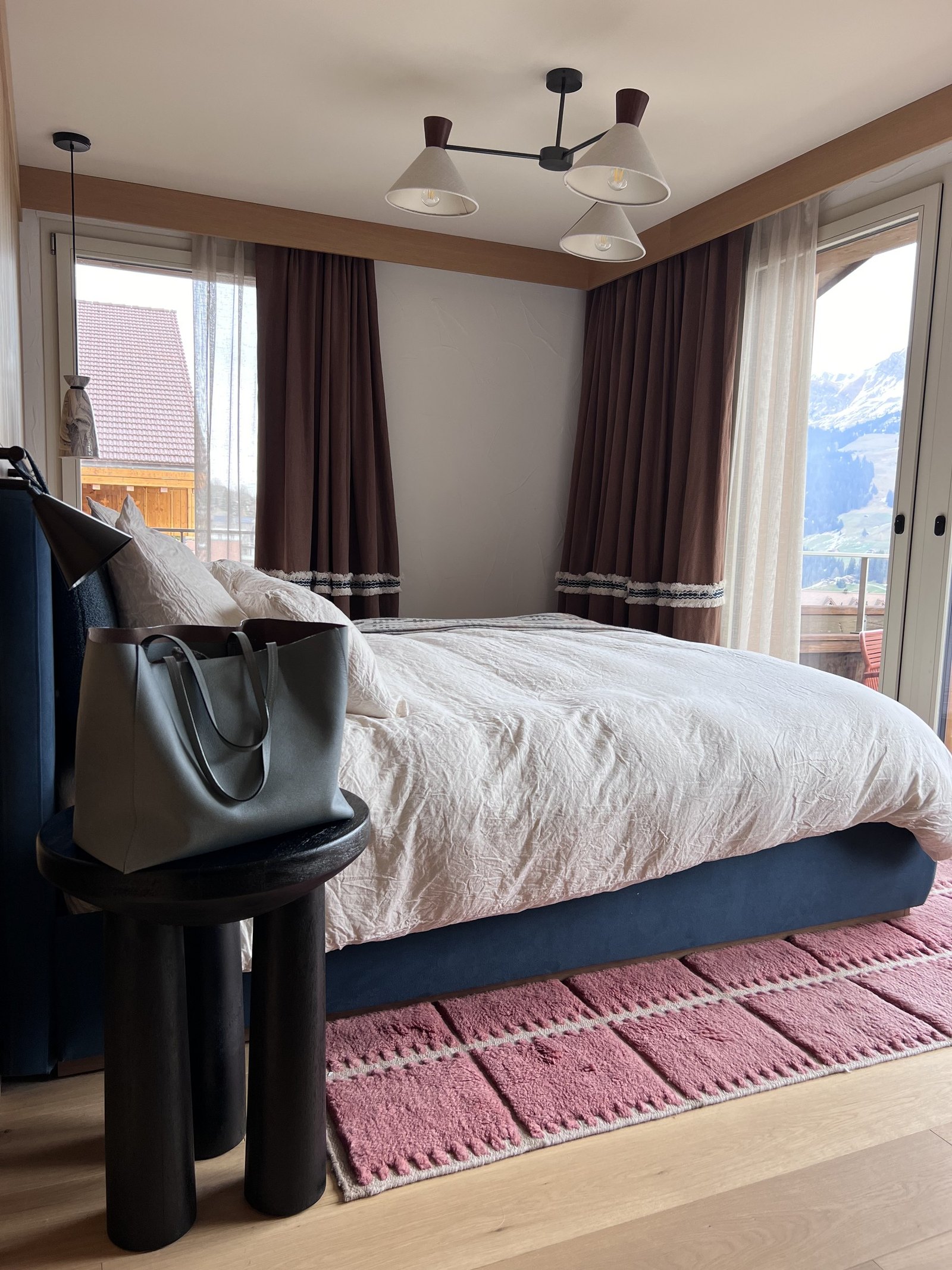 A bed between two windows with brown curtains, a black armchair and a pink striped rug