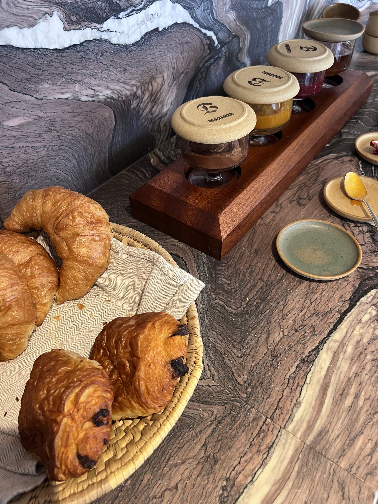 Croissants and pains au chocolat with small jars of jam on a timber rack over marble