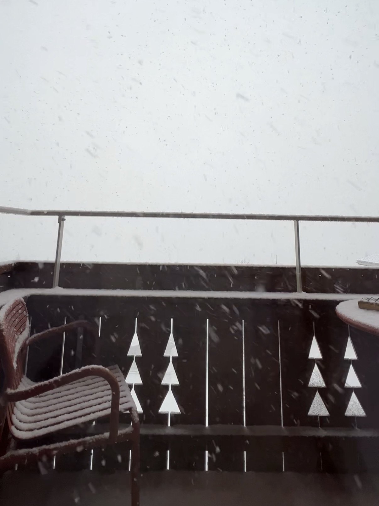 Snow falling past a timber balcony at The Brecon, settling on a red chair and on a balustrade cut with fir tree shapes