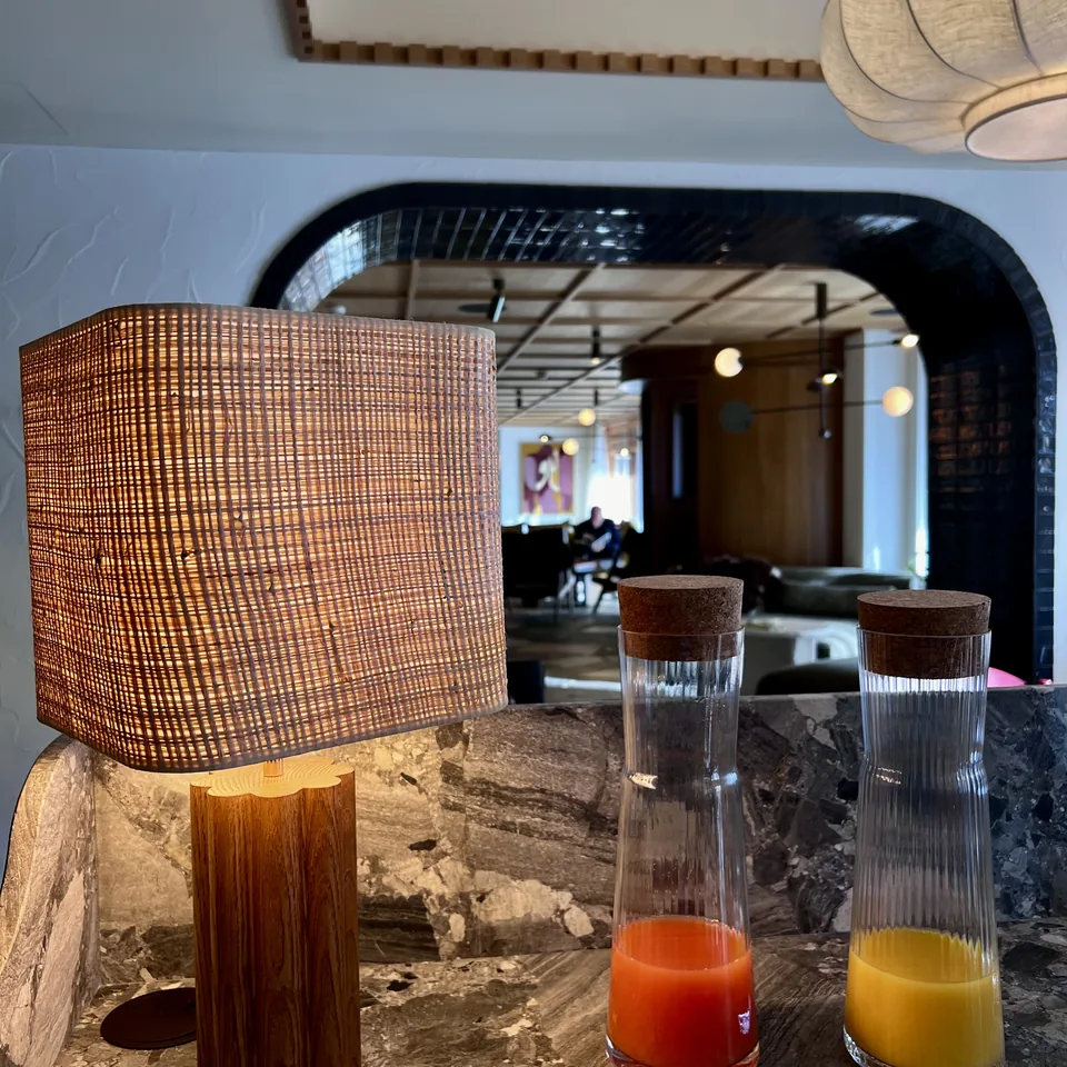 A woven shaded table lamp on a marble counter beside two glass carafes of juice, with the tiled arch beyond
