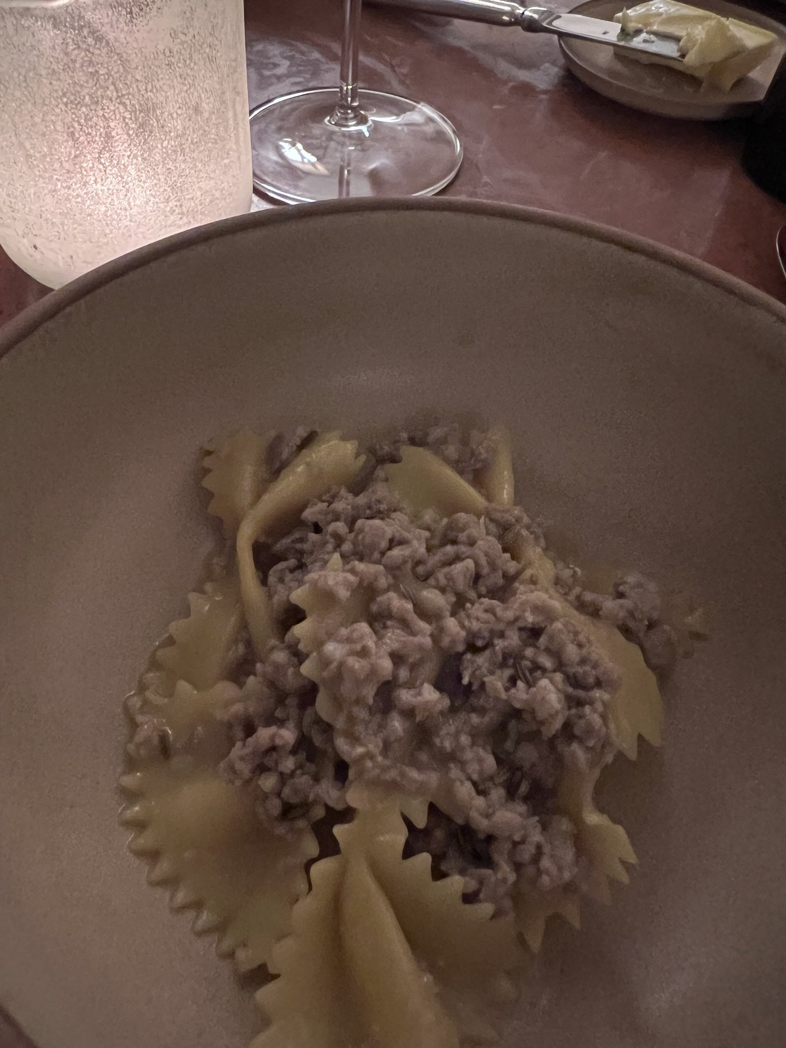 A wide ceramic bowl of ribbon pasta under a meat ragu, with a wine glass behind it