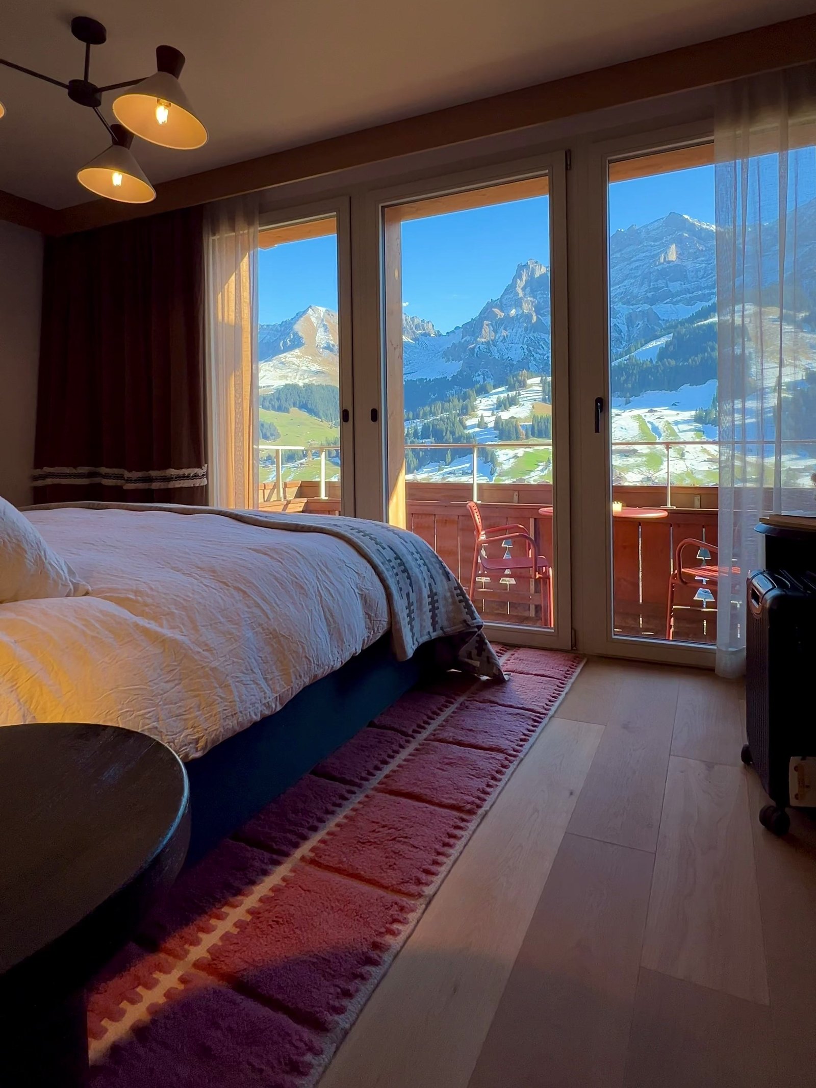 A bedroom at The Brecon in late afternoon light, the glass doors running the width of the wall onto a balcony with red chairs and the snow covered peaks behind