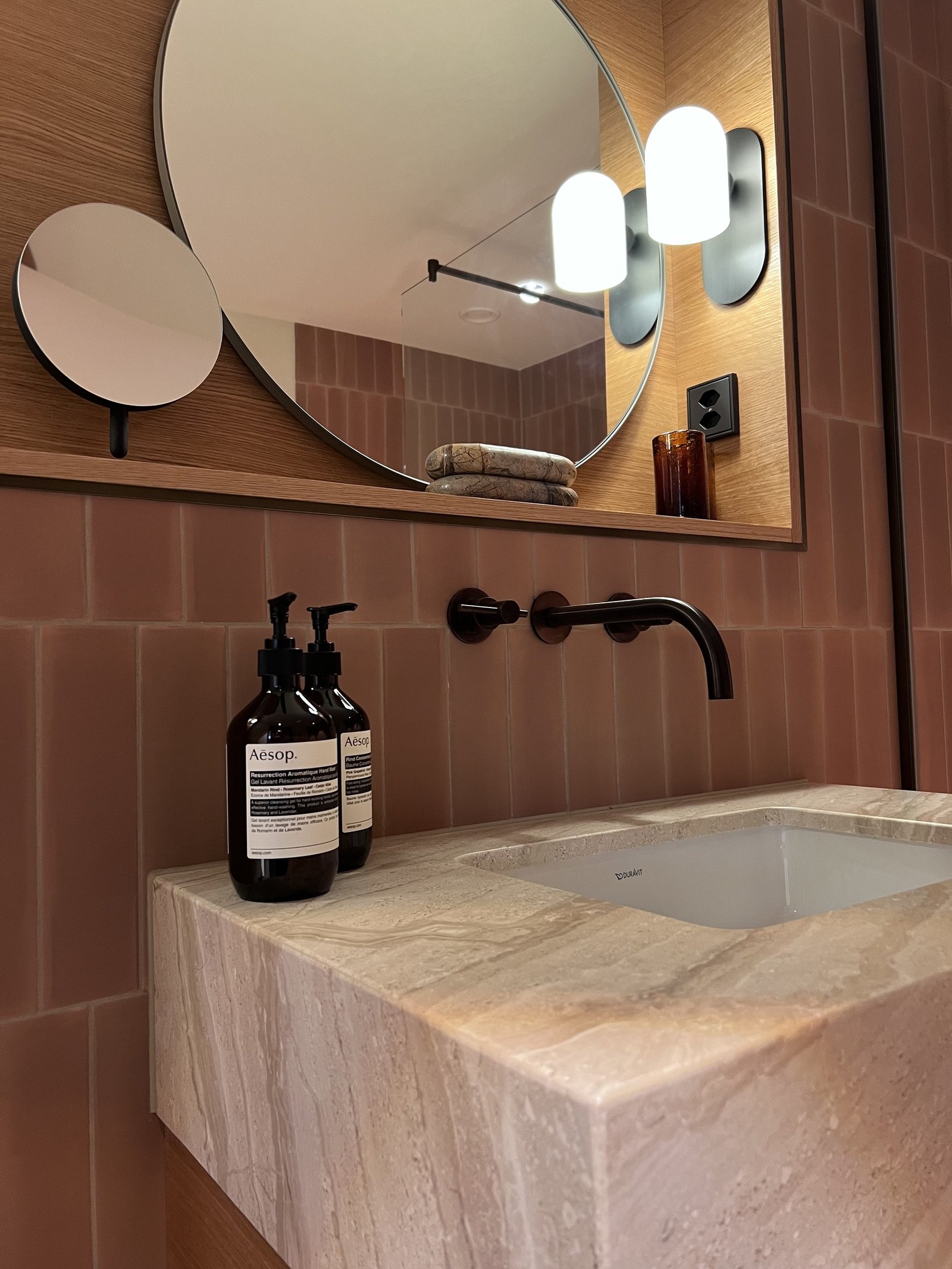A carved stone basin with black taps, Aesop bottles and a round mirror above