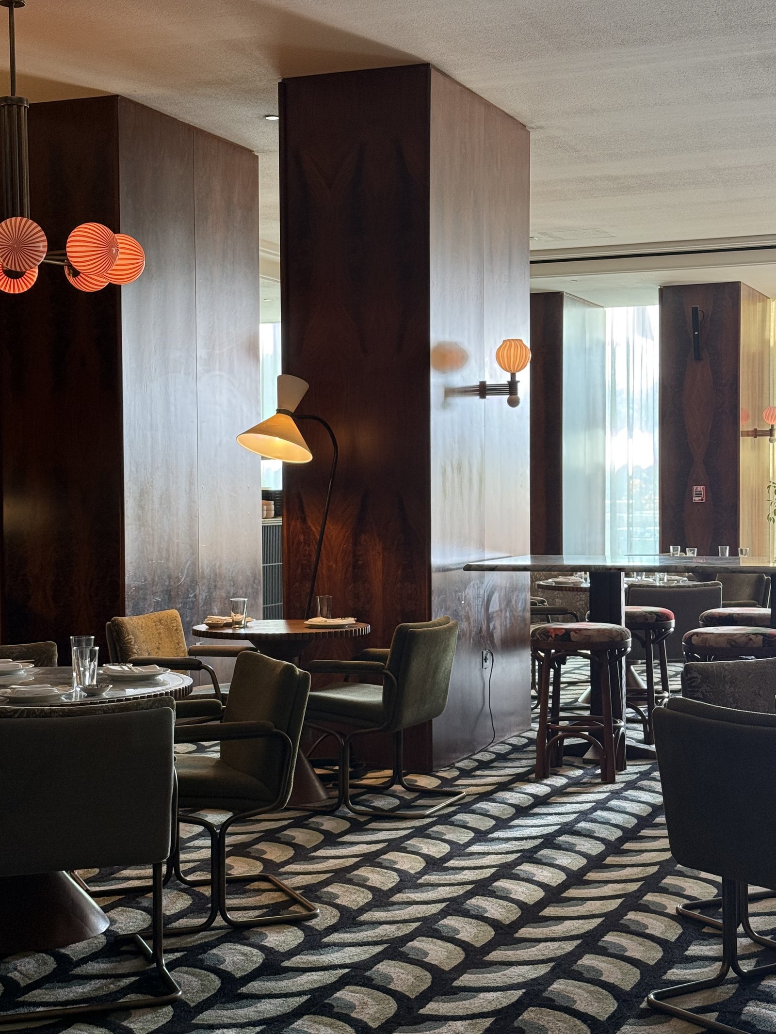 A bar at The Ned Doha with orange globe lights on a brass stem, dark wood columns and a boldly patterned carpet