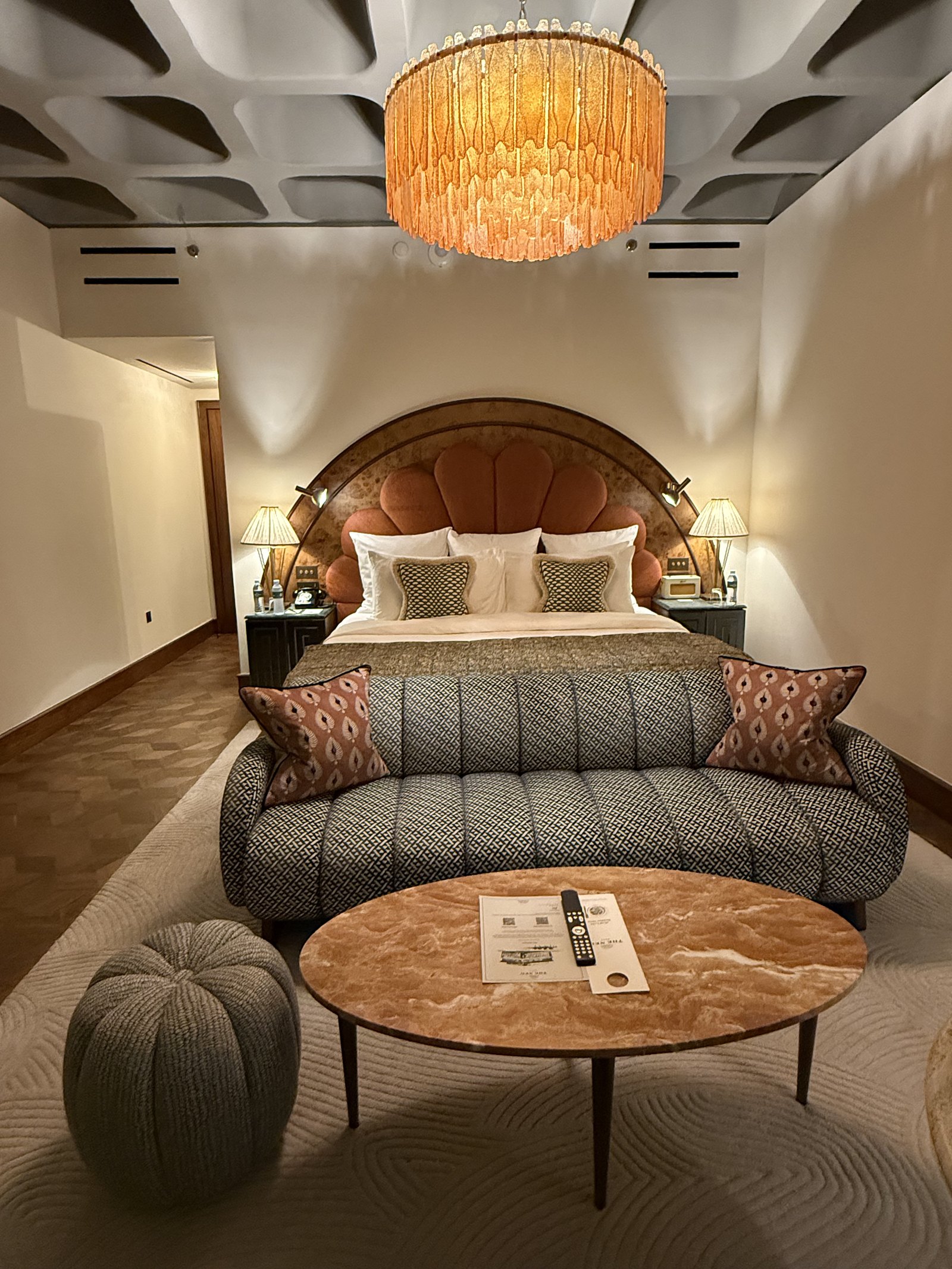 A bedroom at The Ned Doha with a tan velvet shell headboard, a ribbed bed base and a round burl table, under a coffered concrete ceiling and an amber glass chandelier